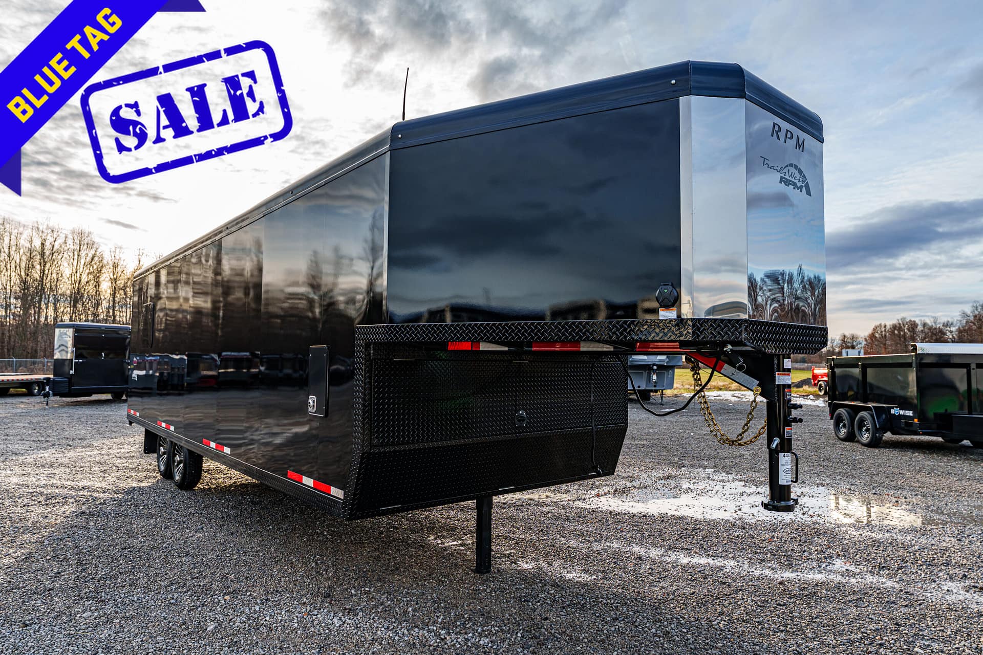 Black enclosed trailer on a gravel lot with a large blue SALE stamp and a blue diagonal BLUE TAG banner in the sky.