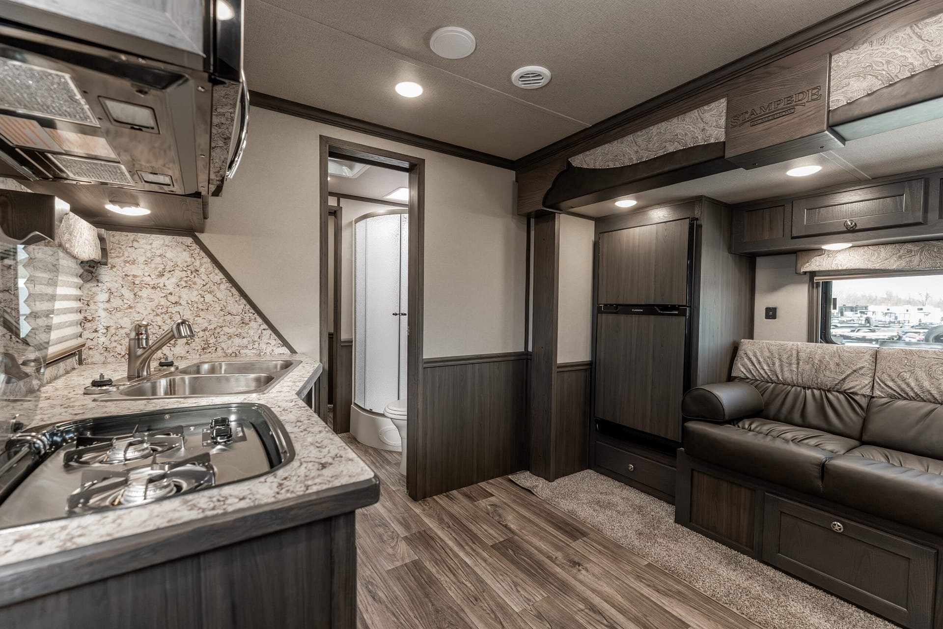 RV kitchenette with gas stove, double sink, granite countertop, and dark wood cabinets; living area to the right.