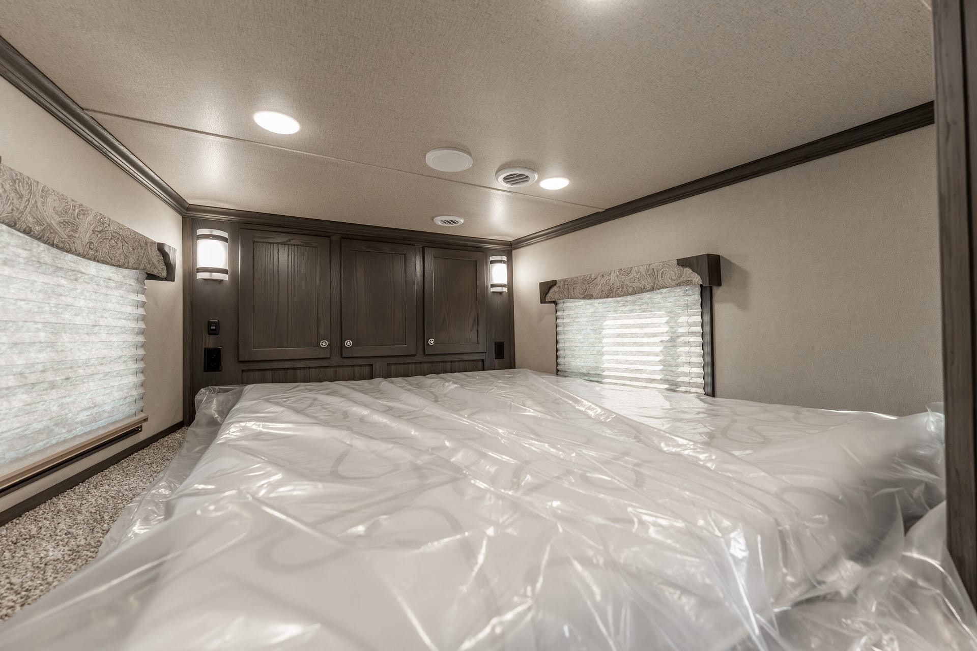 RV bedroom interior with dark wood cabinets above a bed, two wall sconces, and windows with blinds