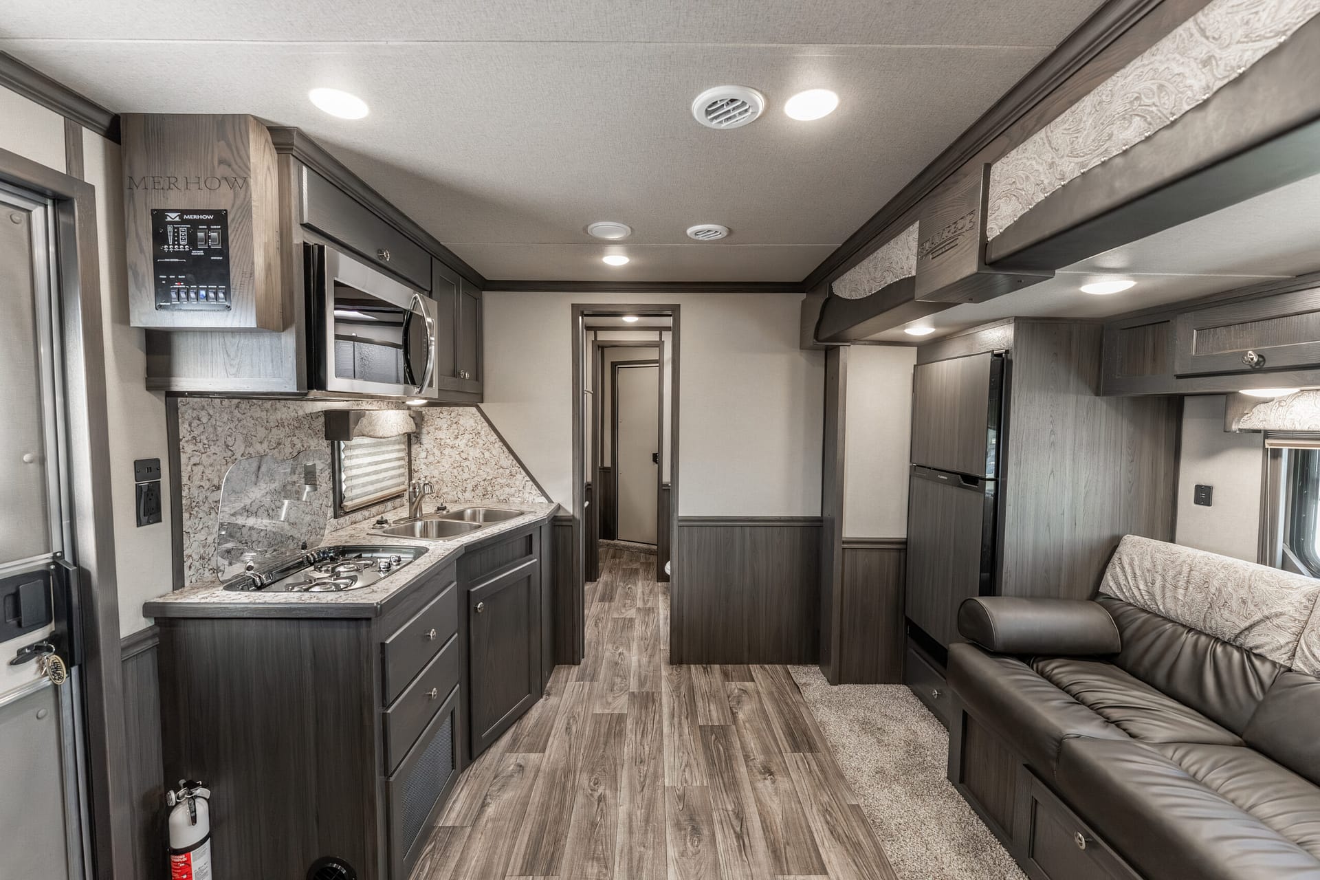 Interior of a modern RV kitchen and living area with dark wood cabinets, stove, microwave, and a sofa along the right wall.