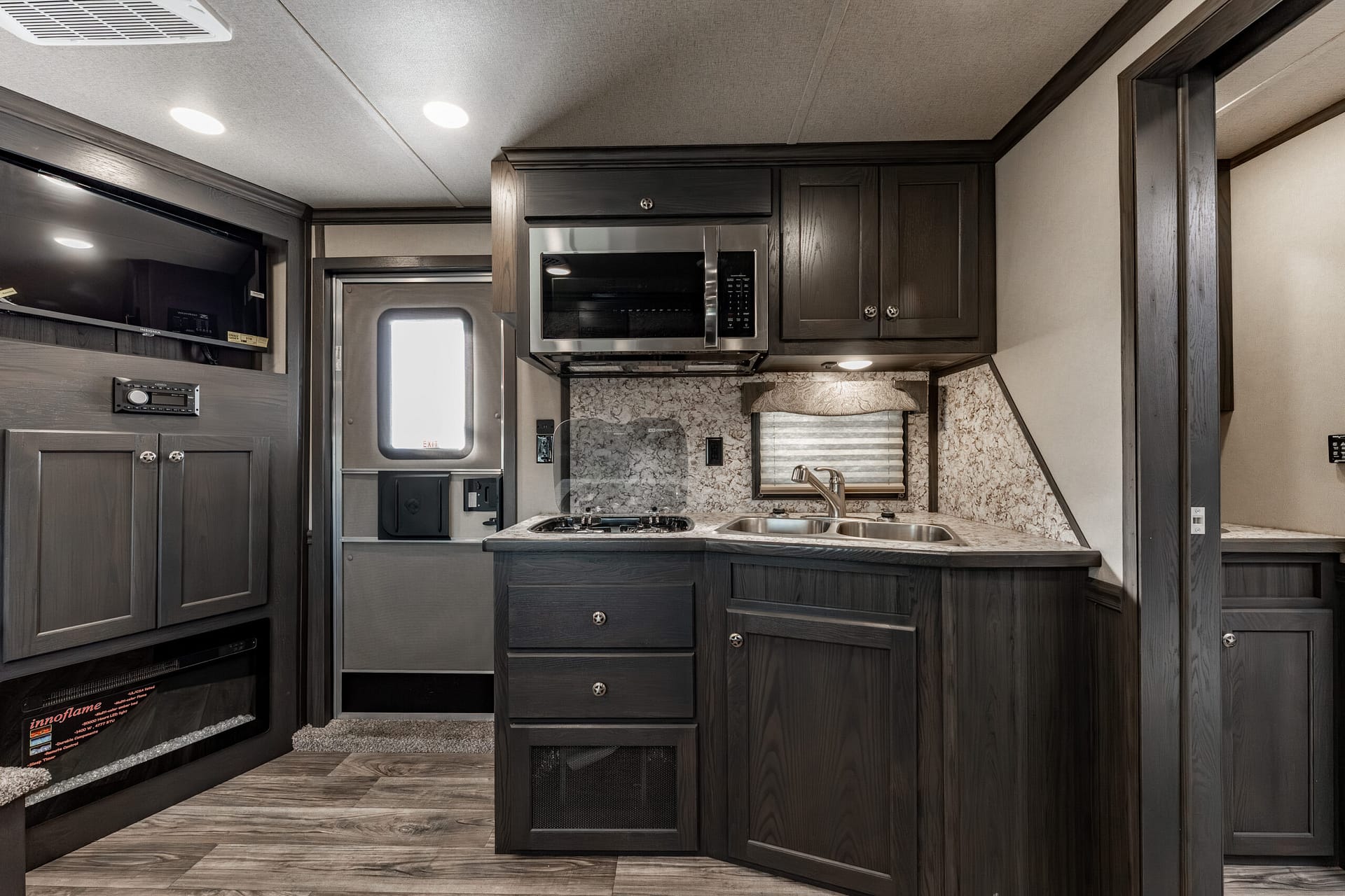 RV kitchen with dark wood cabinets, stainless microwave, stovetop, and double sink on a marble countertop centerpiece