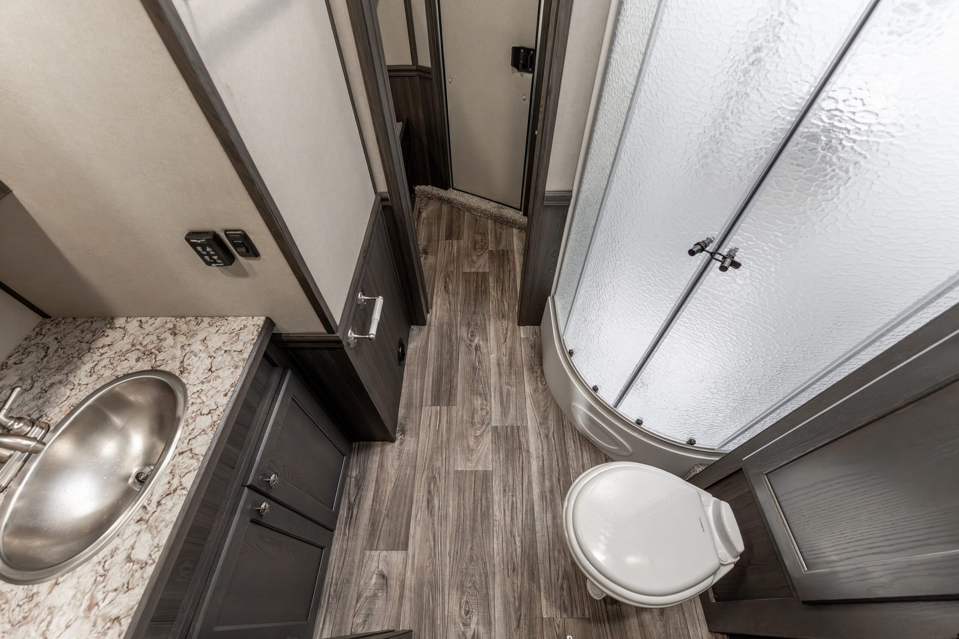 Compact RV bathroom showing a curved glass shower enclosure on the right, a white toilet in the foreground, and a marble countertop with a stainless sink on the left. Dark wood cabinets line the wall; wood-look vinyl flooring runs throughout.