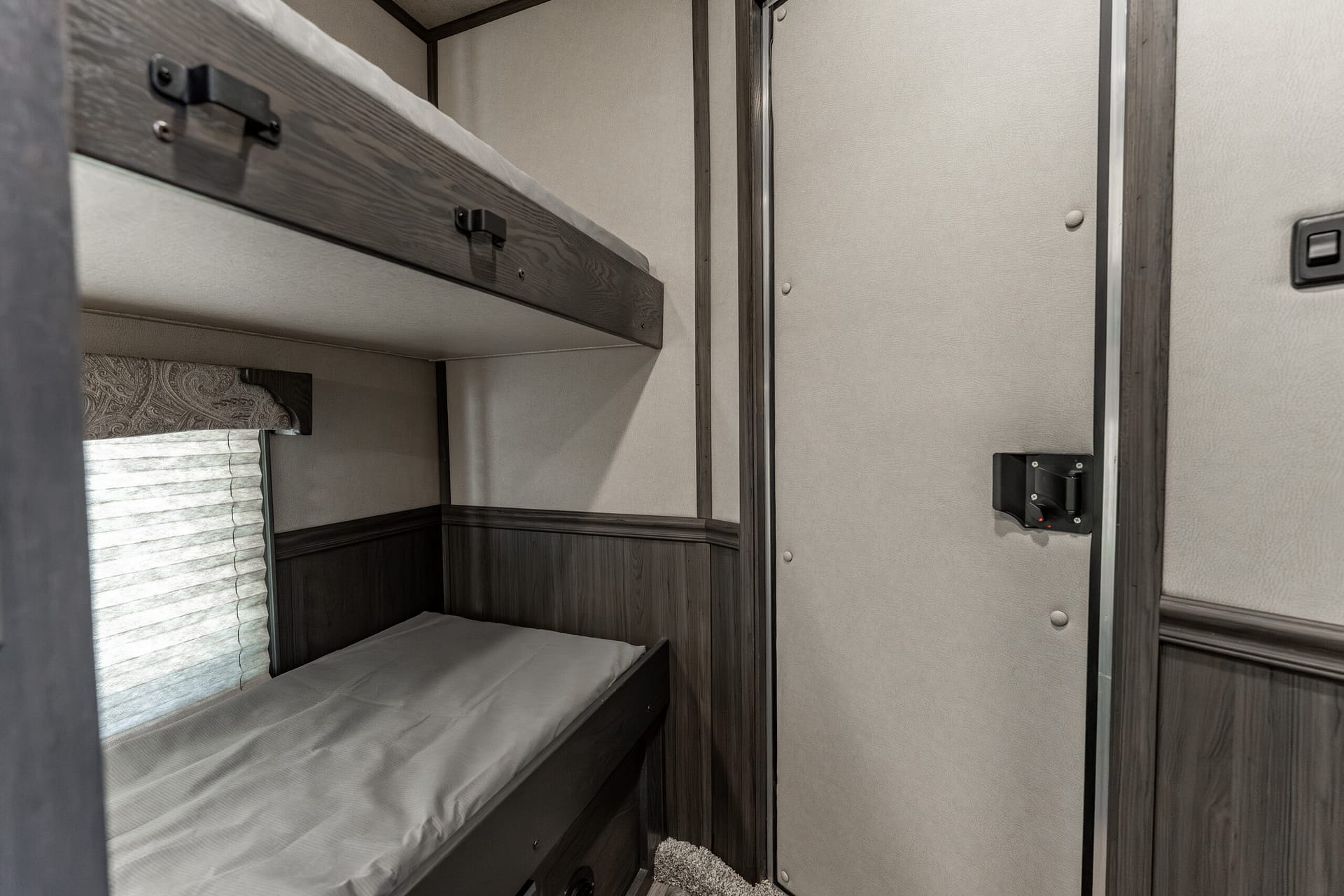Interior of a camper with a lower bunk and an upper bunk, gray bedding and dark wood trim.