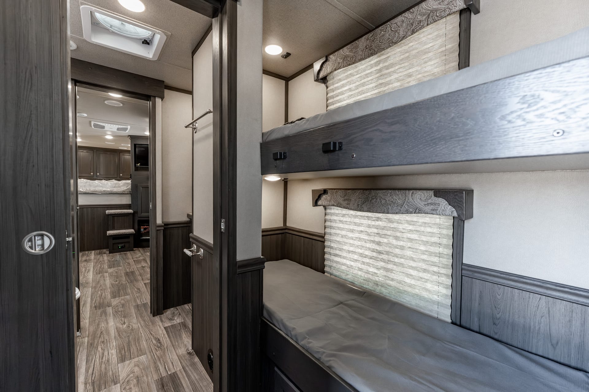 Compact RV bunkroom with two stacked beds, dark wood cabinetry, and striped window shades on the right side