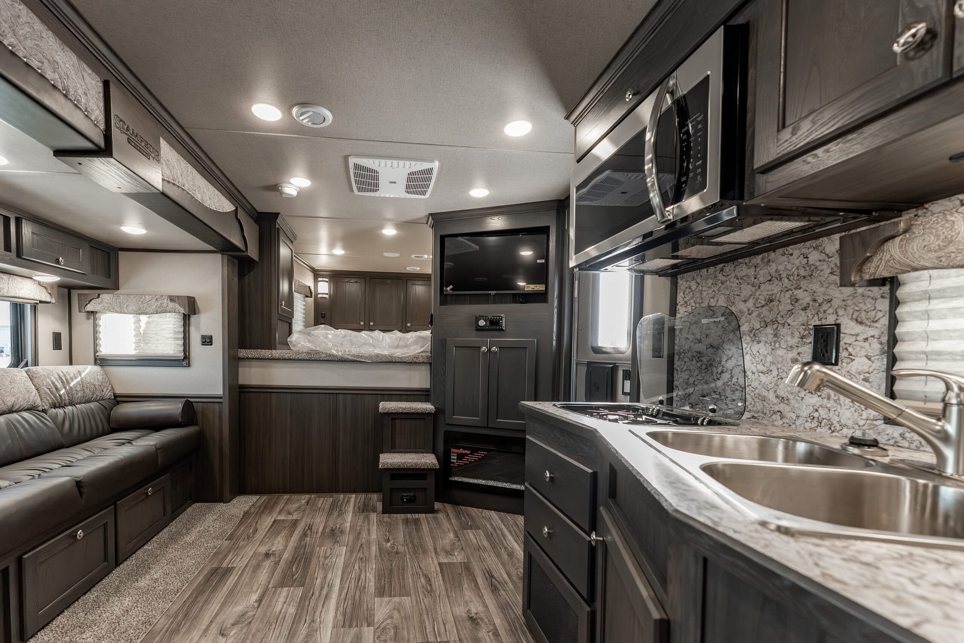 Interior of a dark-wood RV with a long sofa on the left, kitchen island and stainless sink on the right, and overhead cabinets above.”] ,