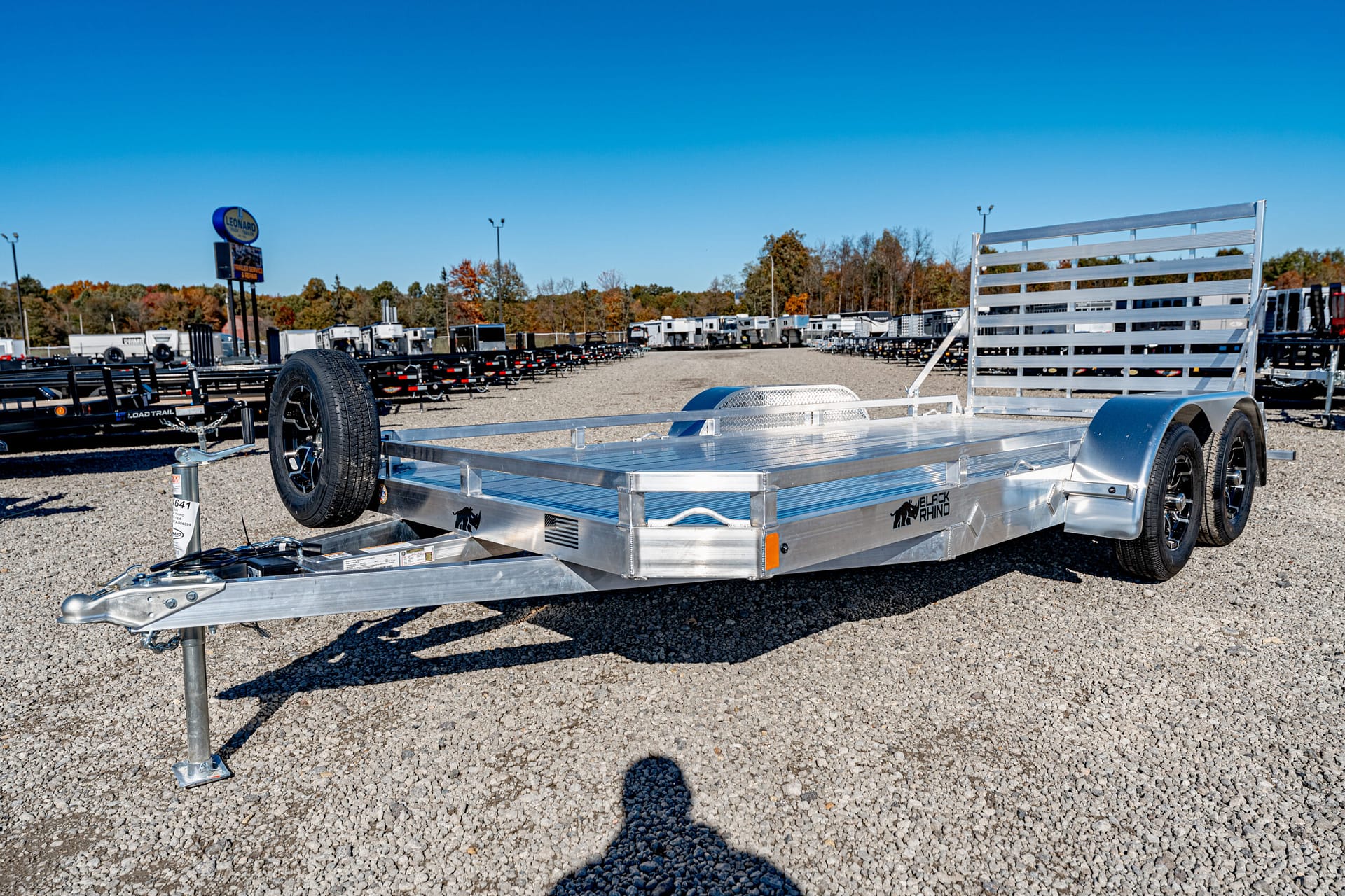 Aluminum flatbed trailer with dual axles and spare tire on a gravel lot at a trailer dealership.