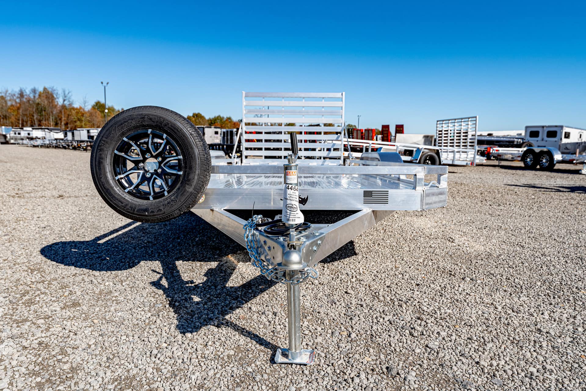 Aluminum utility trailer with a mounted spare tire on the left, safety chains, and a hitch in a gravel lot.