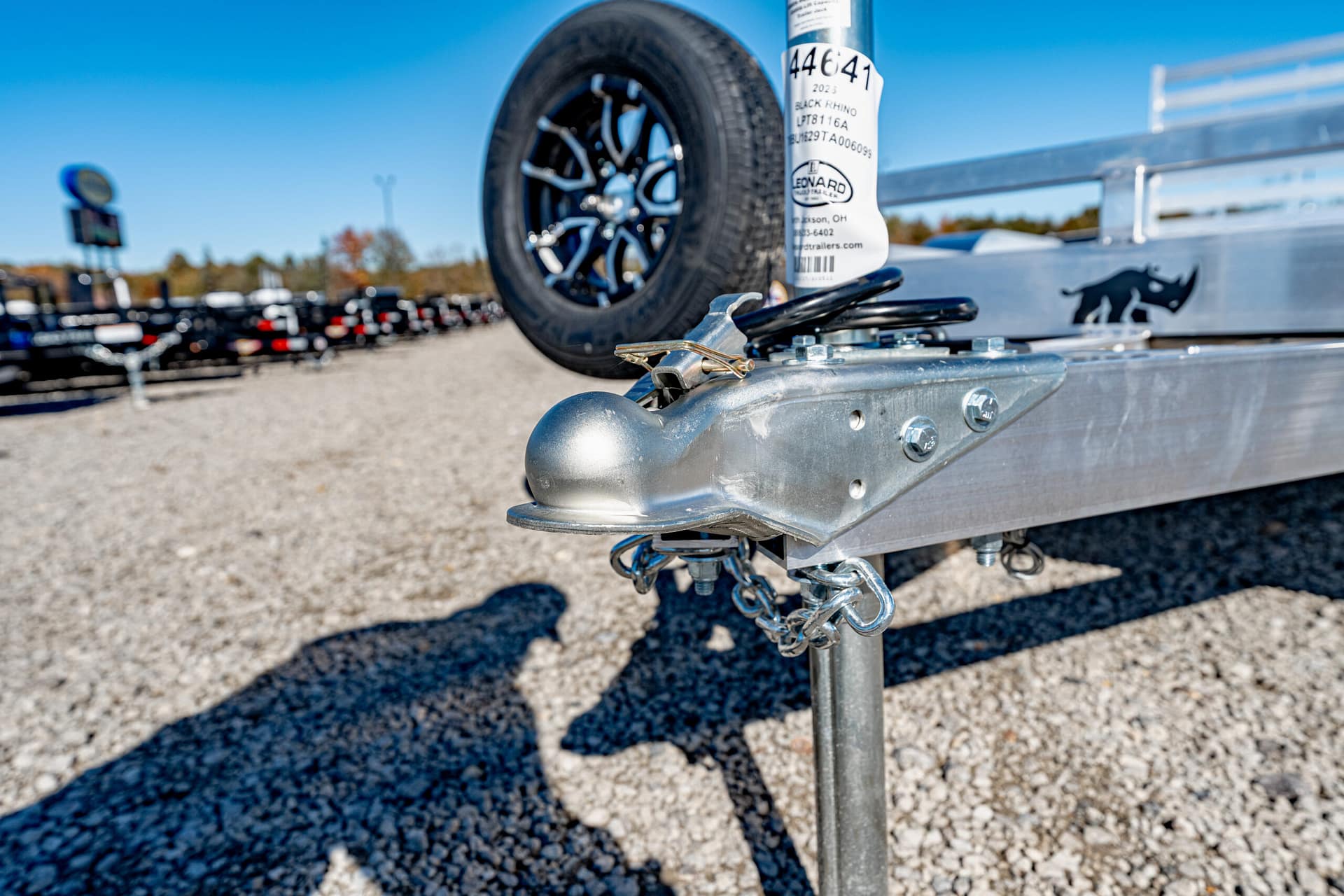 Close-up of a metal trailer hitch with safety chain, mounted on a trailer in a gravel lot.
