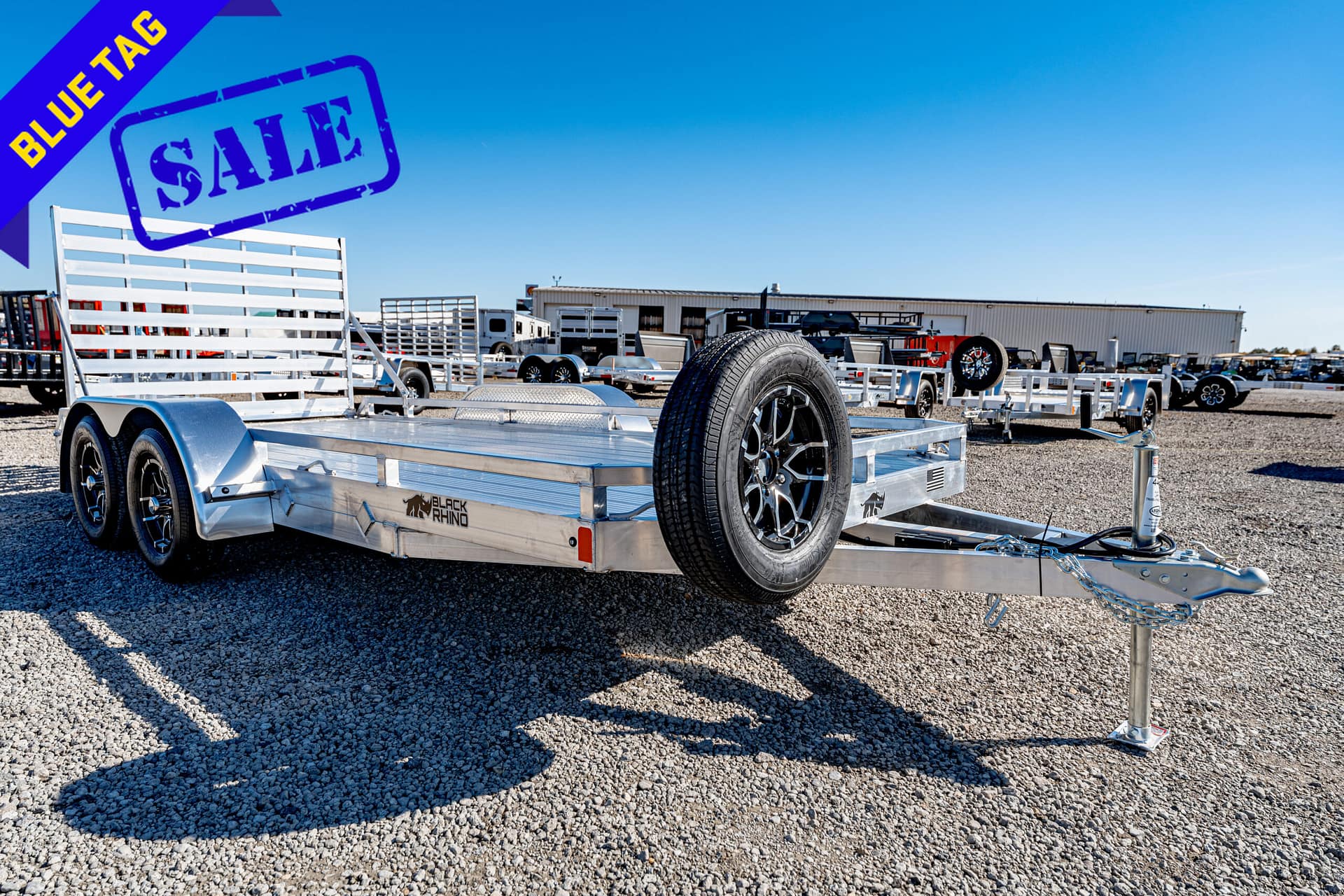 Dual-axle aluminum utility trailer on a gravel lot with a spare tire mounted, and a large blue 'Blue Tag SALE' banner in the corner indicating a sale.
