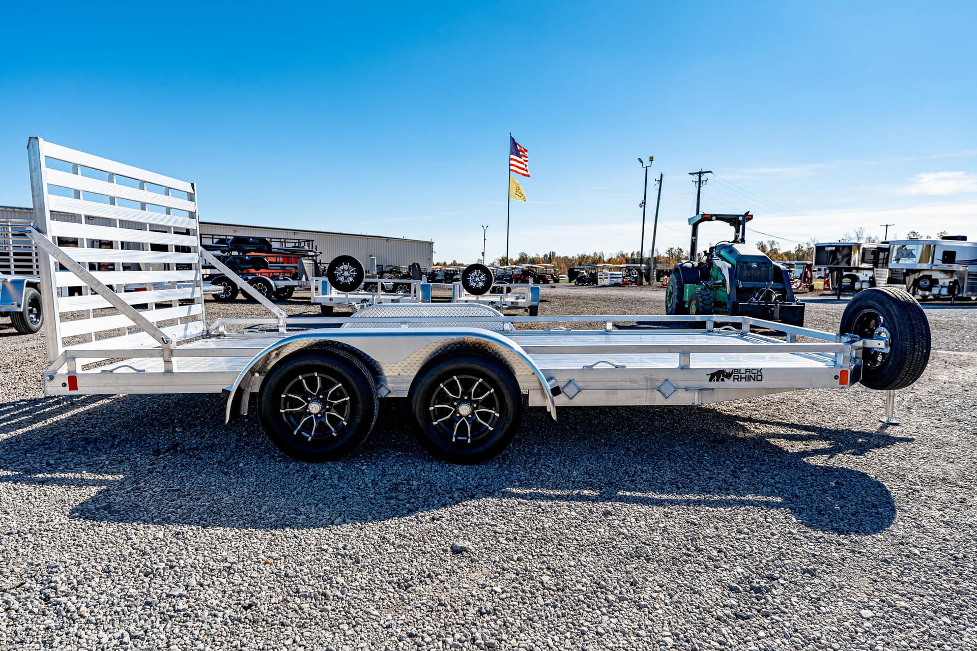 Aluminum dual-axle trailer with a ramp gate on a gravel lot, spare wheel mounted, and a Black Rhino logo visible under bright blue sky