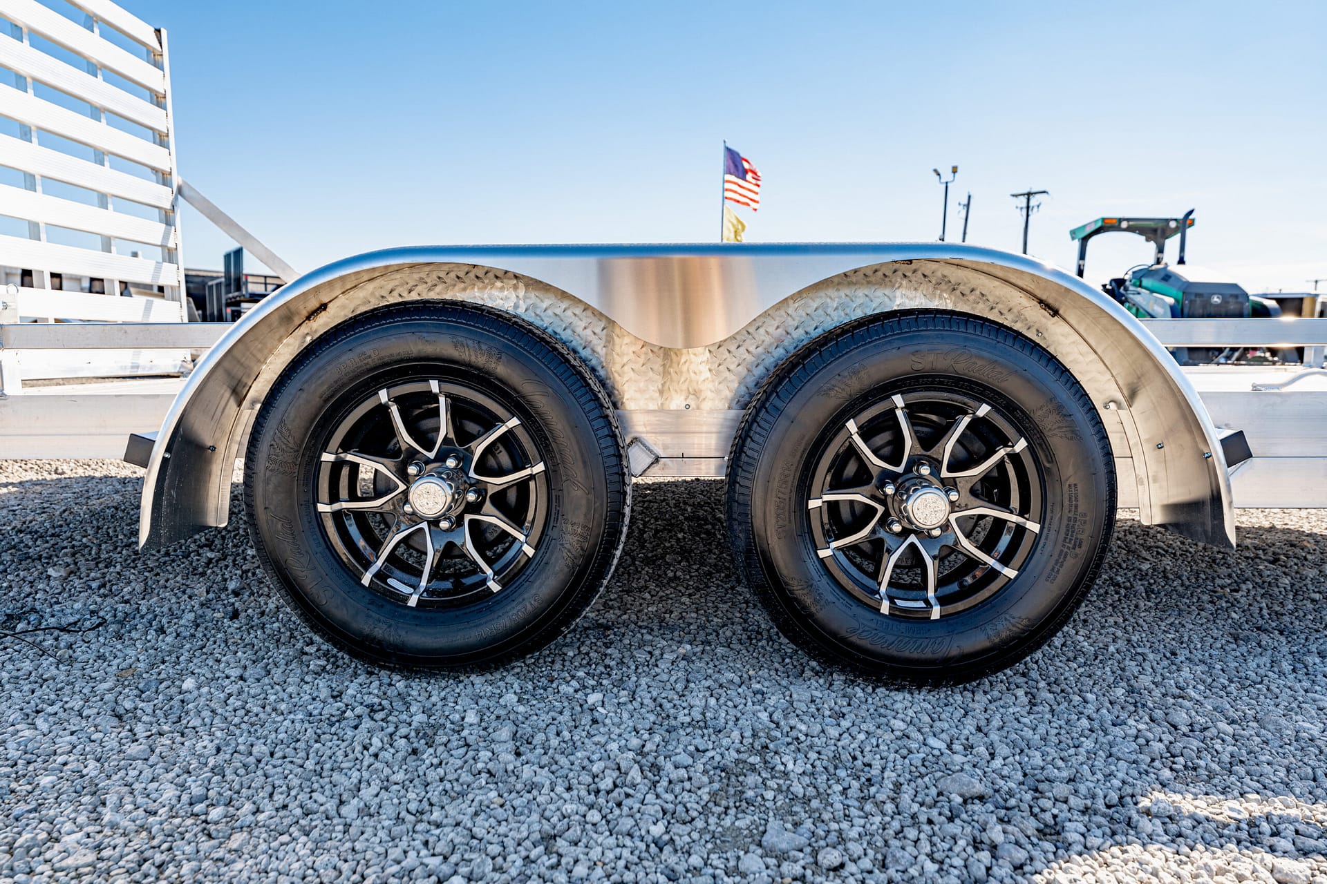 Twin-axle trailer wheels with black and chrome rims on a gravel surface, aluminum fenders above.