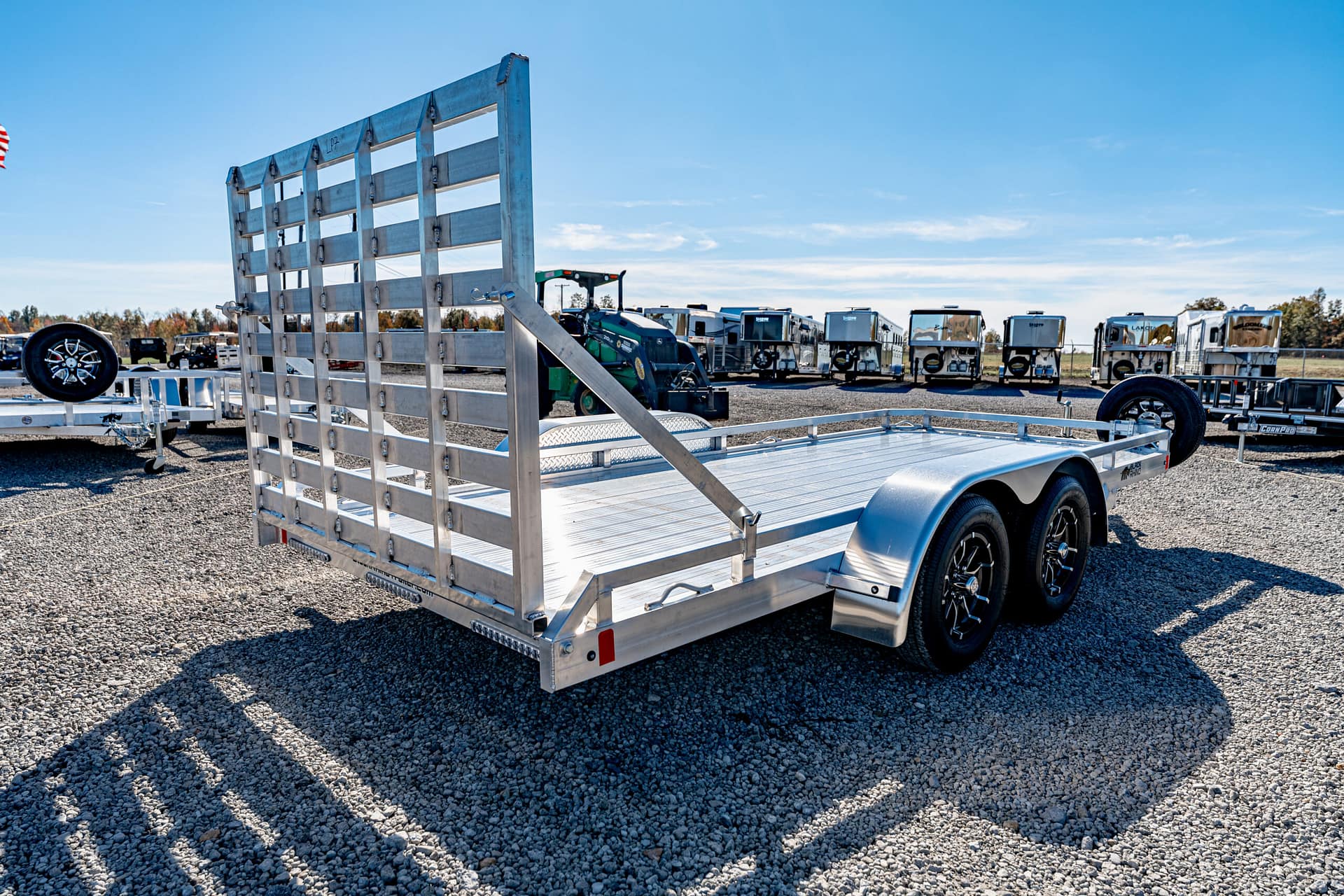 Aluminum flatbed trailer with a tall slatted ramp gate in a gravel lot, surrounded by other trailers under a blue sky.