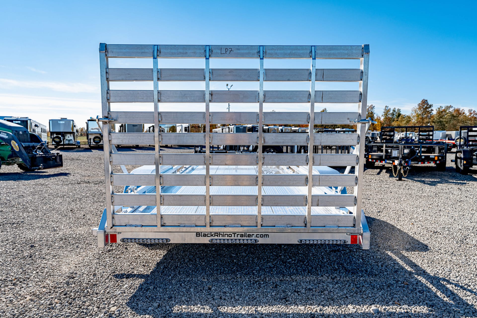 Rear view of an aluminum flatbed trailer with vertical railing slats, parked on a gravel lot; BlackRhinoTrailer.com branding visible on the tailgate.