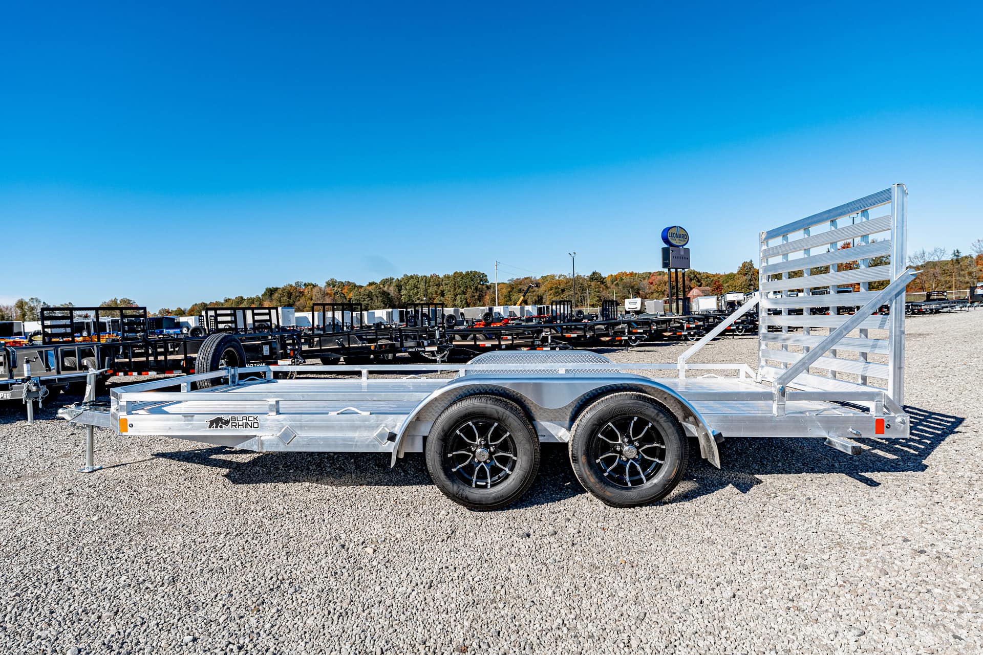 Aluminum flatbed trailer with dual axles and a rear fold-up ramp gate on a gravel lot, surrounded by other trailers at a dealership.