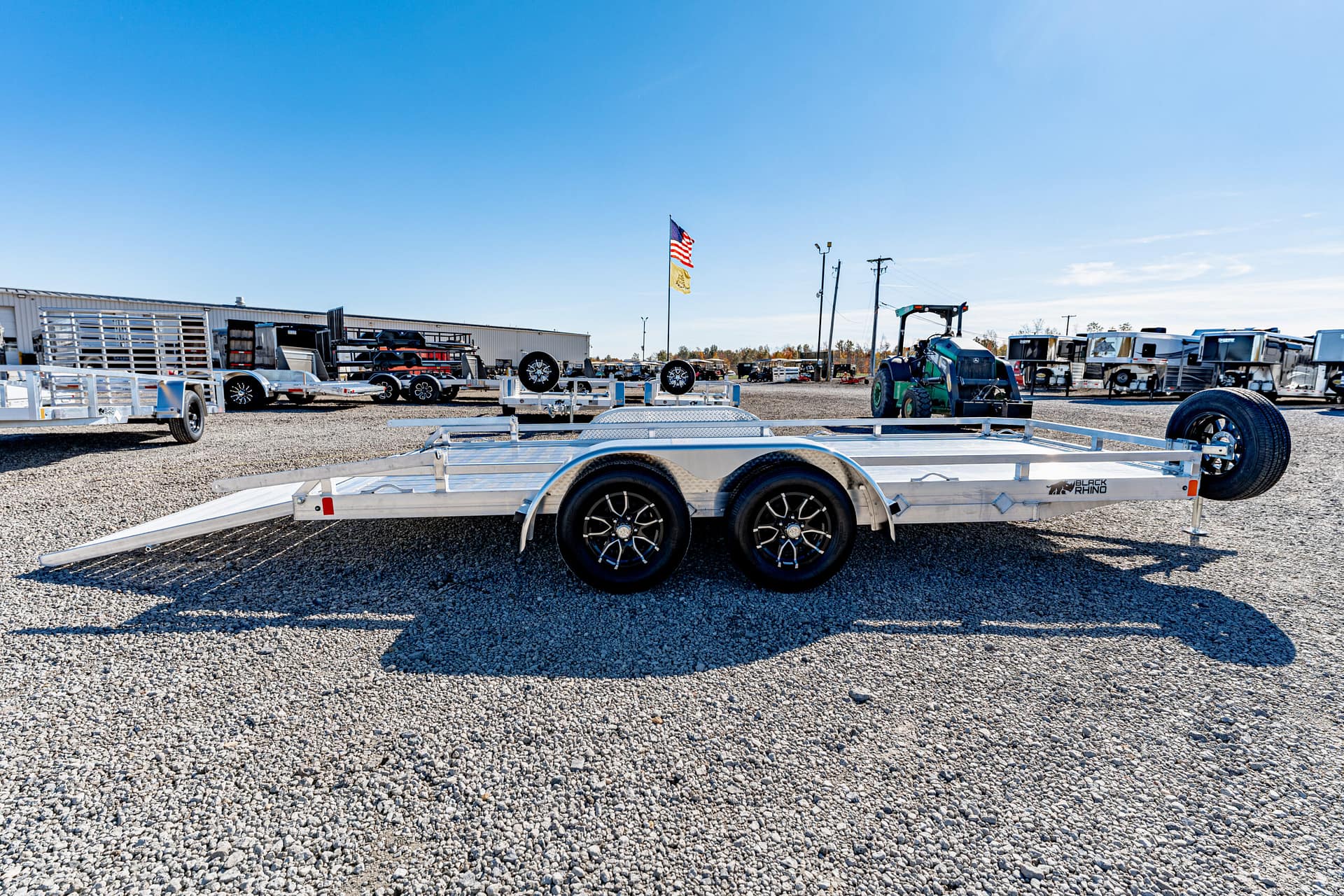 Aluminum car hauler trailer with tandem axles and ramps down, parked in a gravel trailer yard with other trailers and equipment in the background.