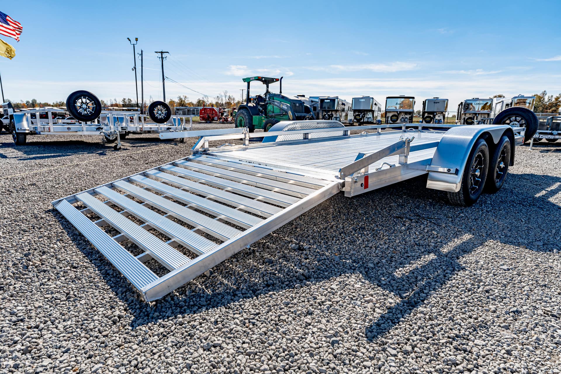 Aluminum car hauler trailer with its loading ramp extended on a gravel lot, other trailers visible in the background.