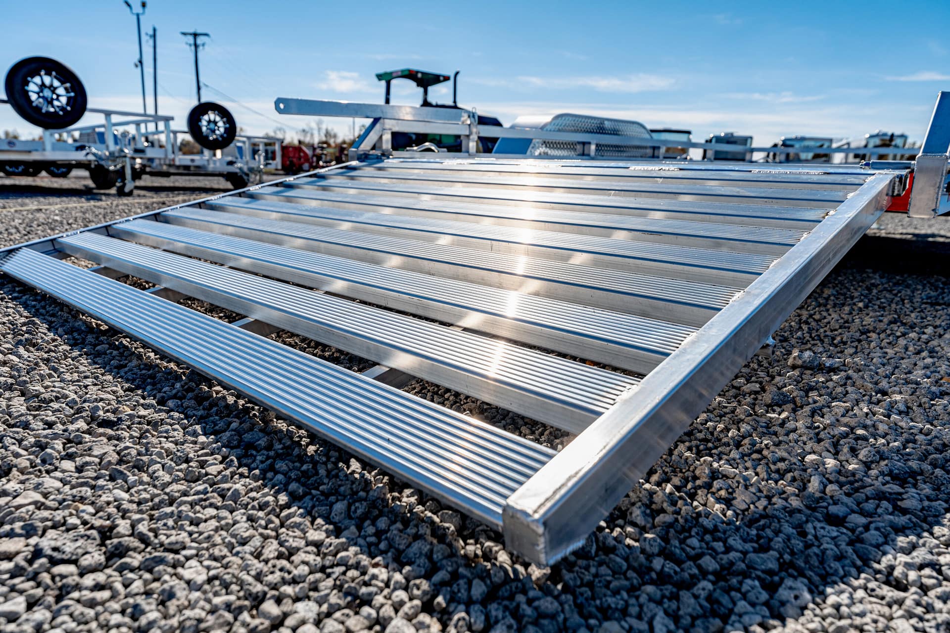 Angle view of a ribbed aluminum trailer ramp resting on a gravel lot with trailers in the background at a storage yard.