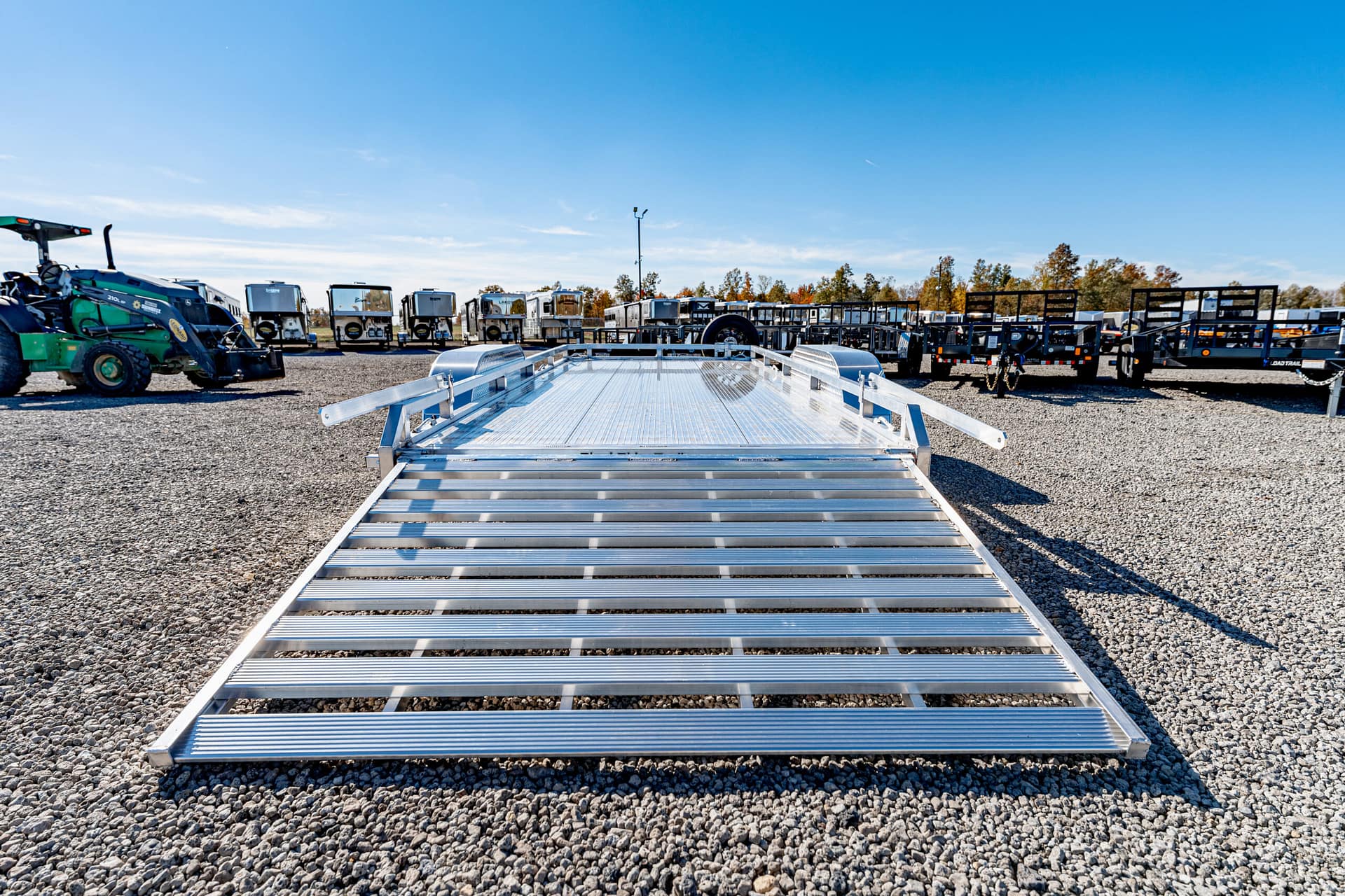 Silver aluminum flatbed trailer with grated ramp, parked on a gravel lot with other trailers in the background.