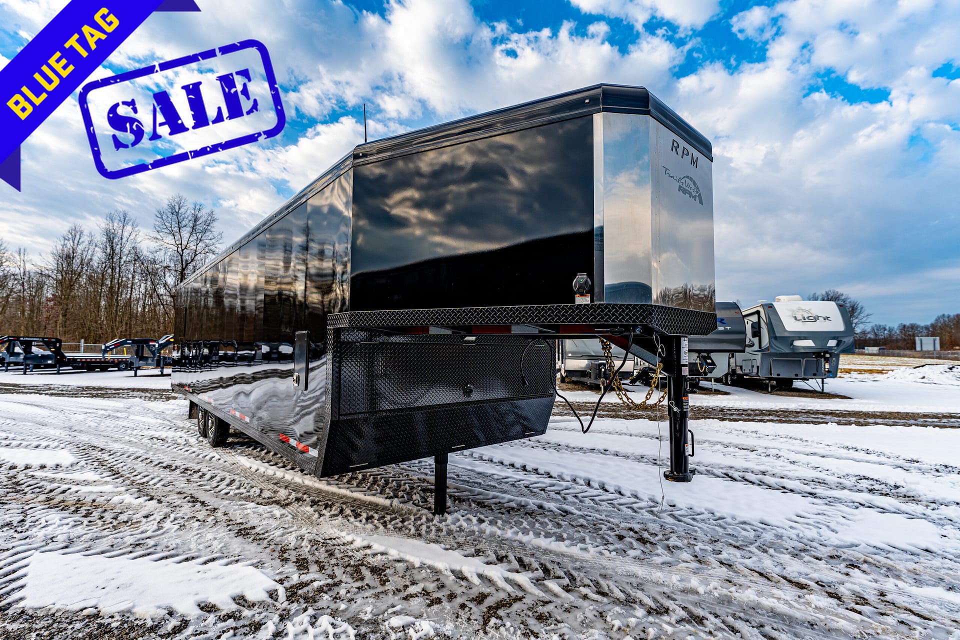 Shiny black enclosed trailer in a snowy lot with a bold blue sale stamp overlapping a purple-blue 'BLUE TAG' ribbon in the sky above it.