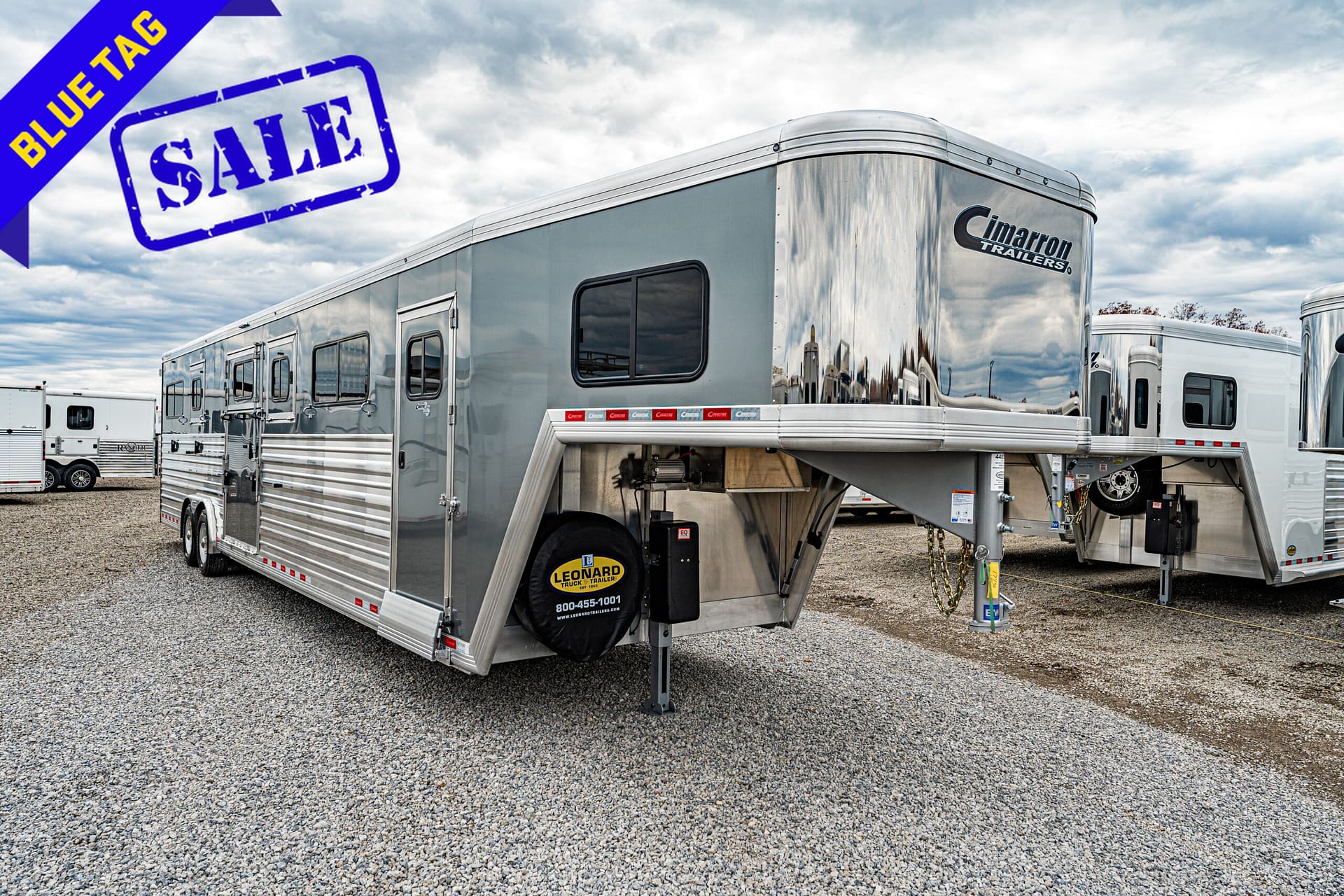 Silver Cimarron trailer on a gravel lot with a large blue SALE stamp and a blue 'Blue Tag' banner overhead in the foreground.