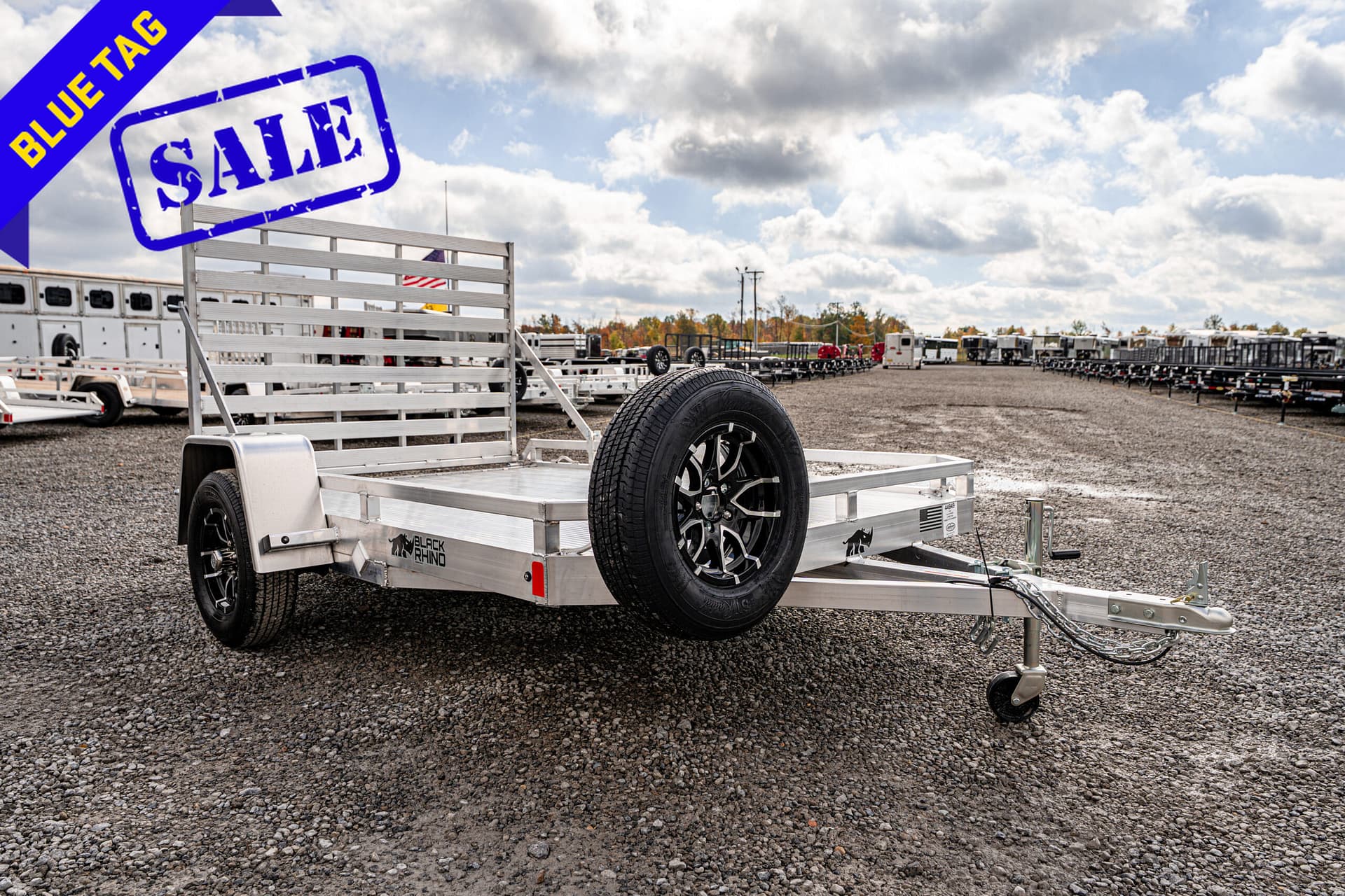 Aluminum utility trailer with a spare tire, parked on a gravel trailer lot; a blue 'BLUE TAG SALE' banner overlays the image.