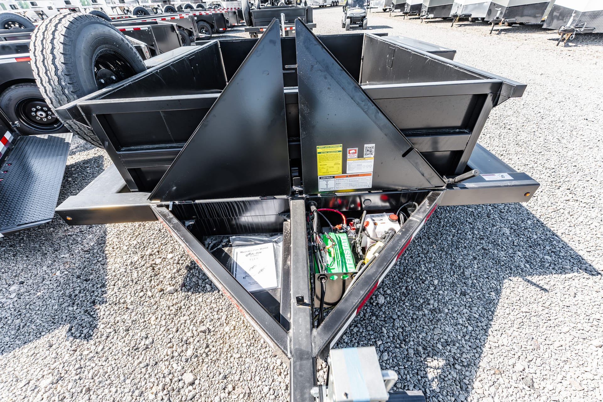 Close-up of a black dump trailer frame with hydraulic mechanism, spare tire, and warning labels on a gravel lot with other trailers nearby