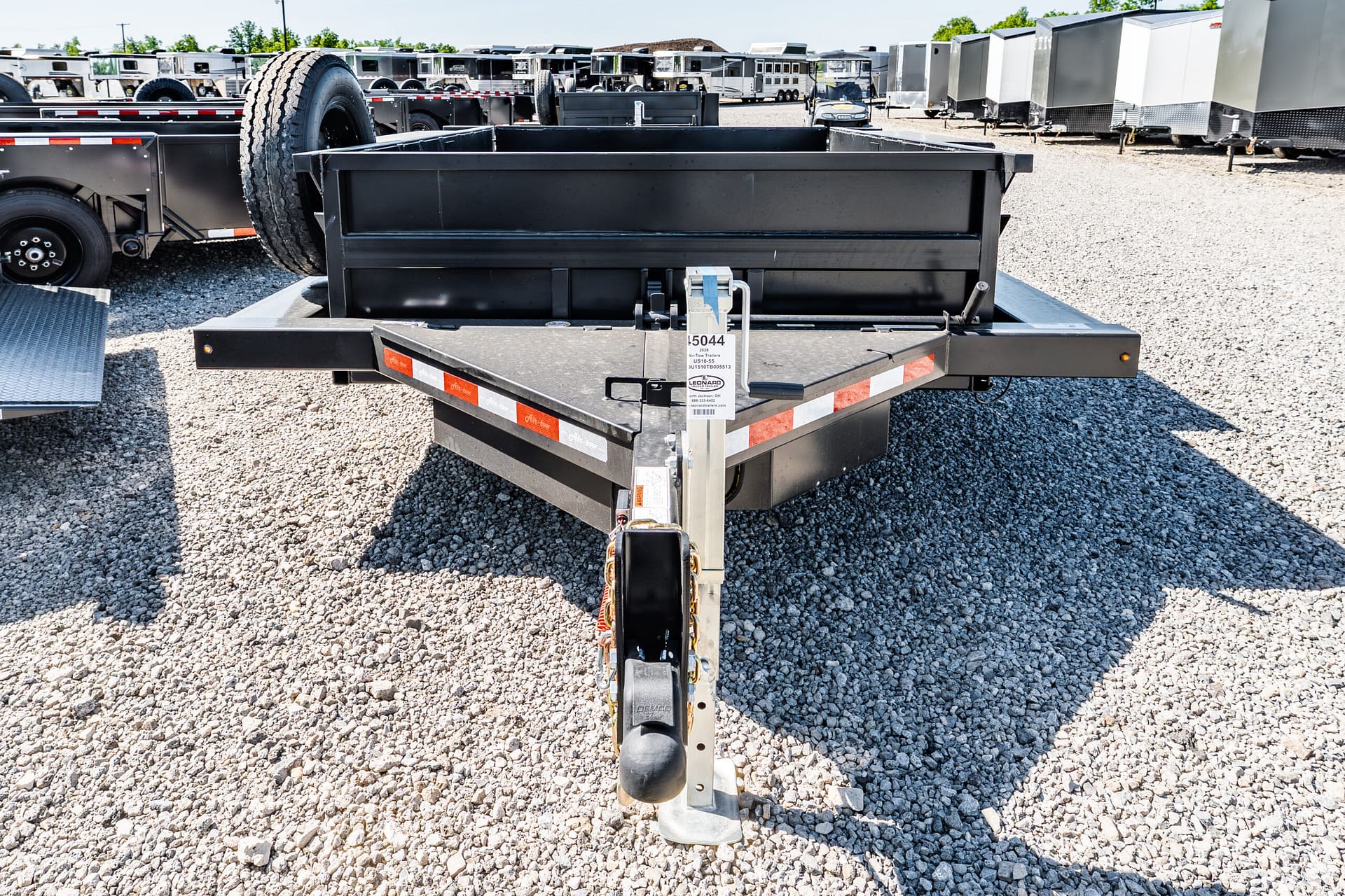Close-up of a black trailer hitch on a gravel lot, with a spare tire and rows of trailers in the background.