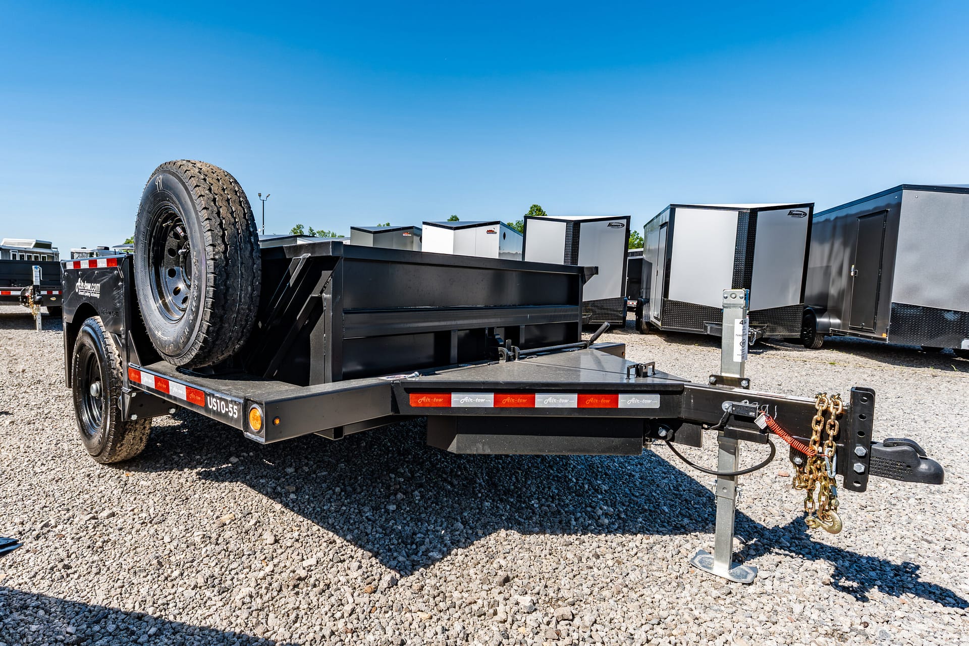 Trailer with a large spare tire mounted on the bed, in a gravel lot with several enclosed cargo trailers under a clear blue sky