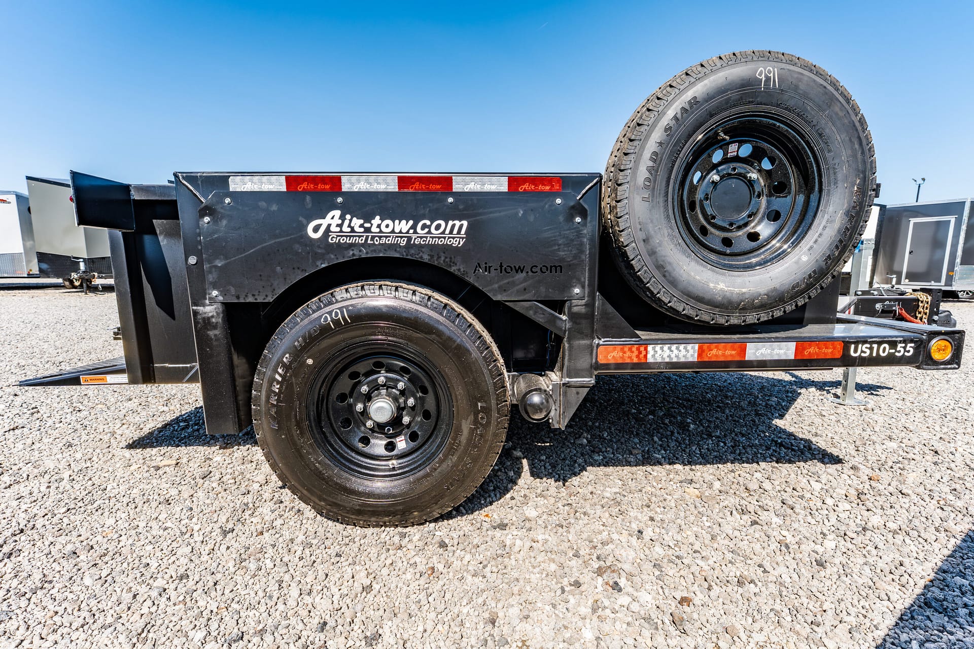 Black trailer with a large spare wheel mounted on the side, Air-tow branding, on a gravel lot under a blue sky.