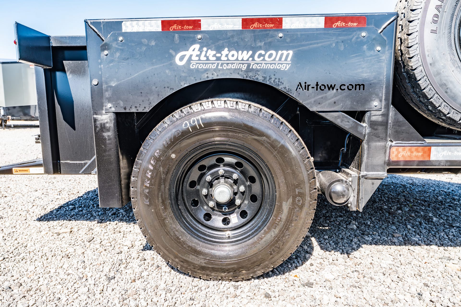 Close-up of a heavy-duty trailer wheel and black metal fender with Air-tow branding and a spare tire mounted on the side, gravel ground beneath.