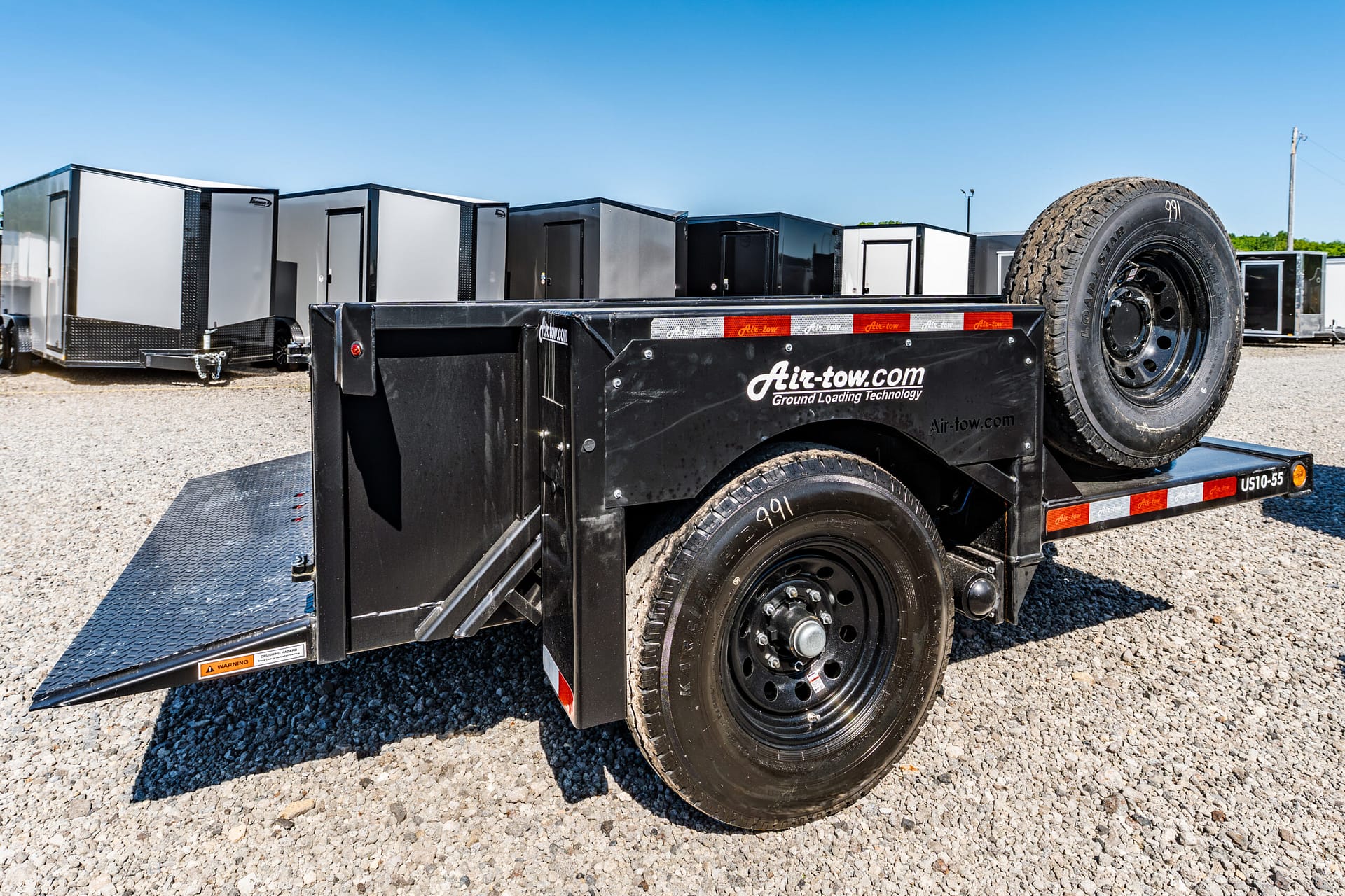 Black utility trailer with a mounted spare tire on the front, parked on gravel, with storage trailers in the background under a clear sky.