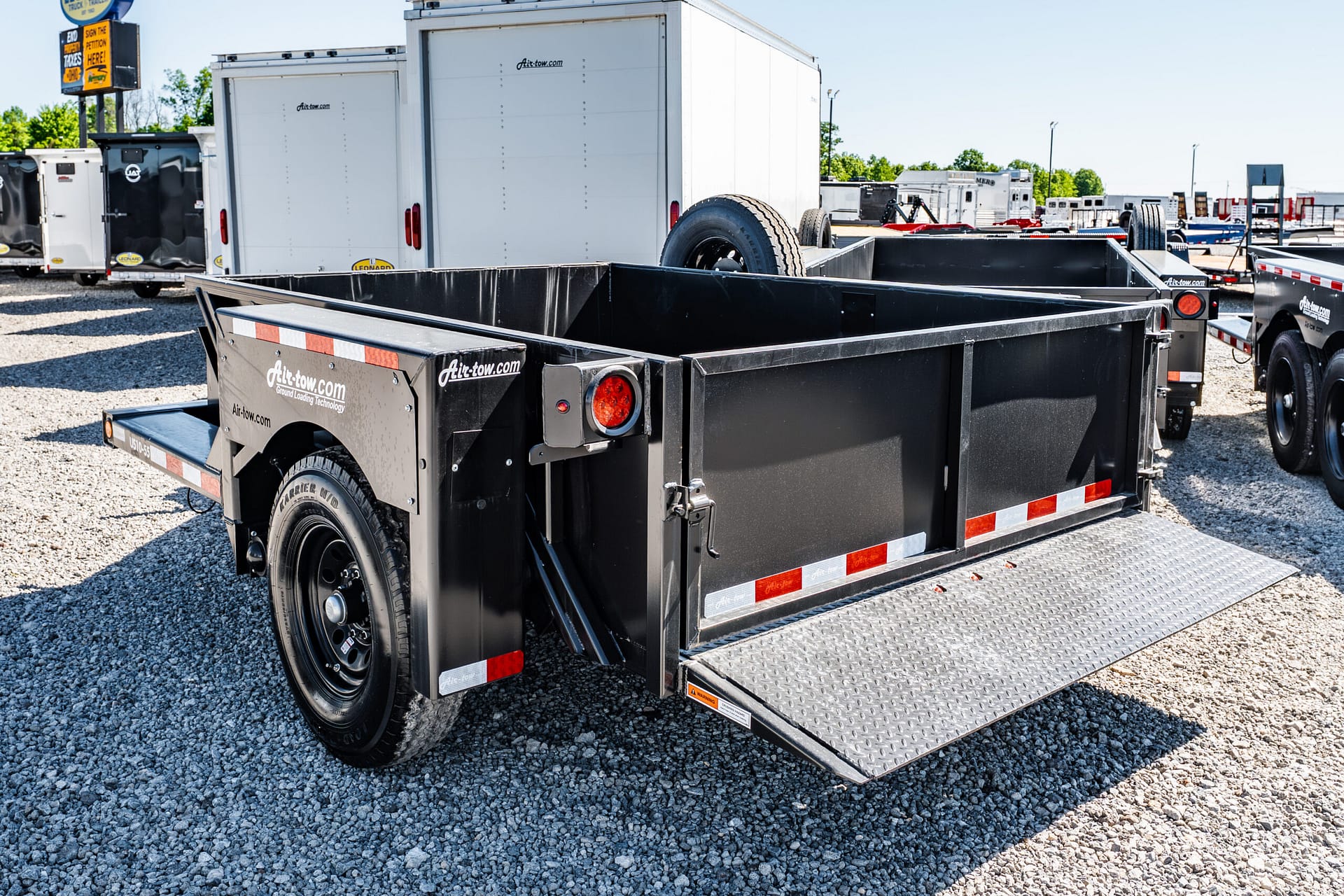 Black utility trailer with its ramp down in a gravel lot, surrounded by other trailers in a trucking yard.