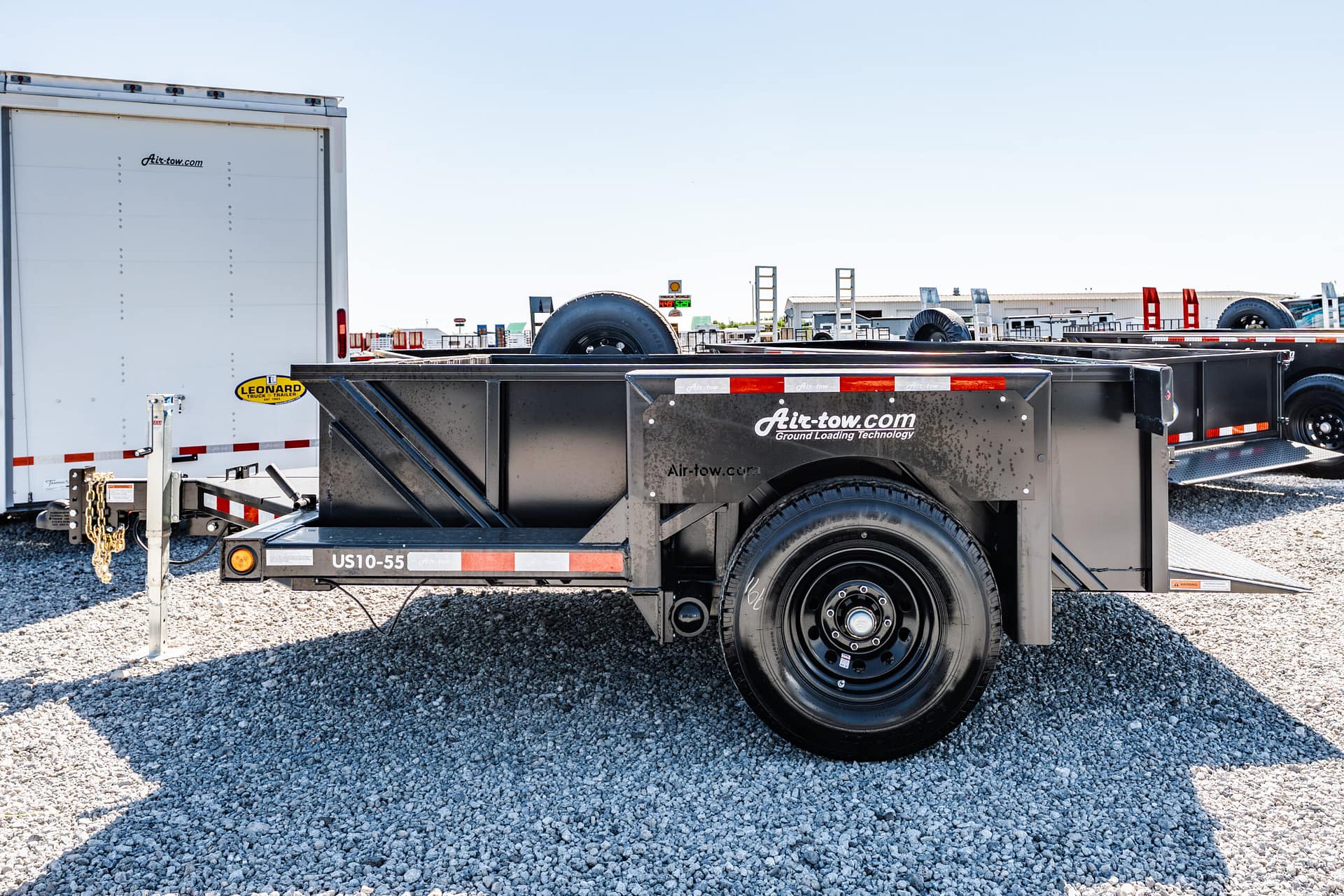 Black Air-tow ground-loading trailer with spare tire and red-and-white reflective stripes on a gravel lot, adjacent to a white trailer truck.