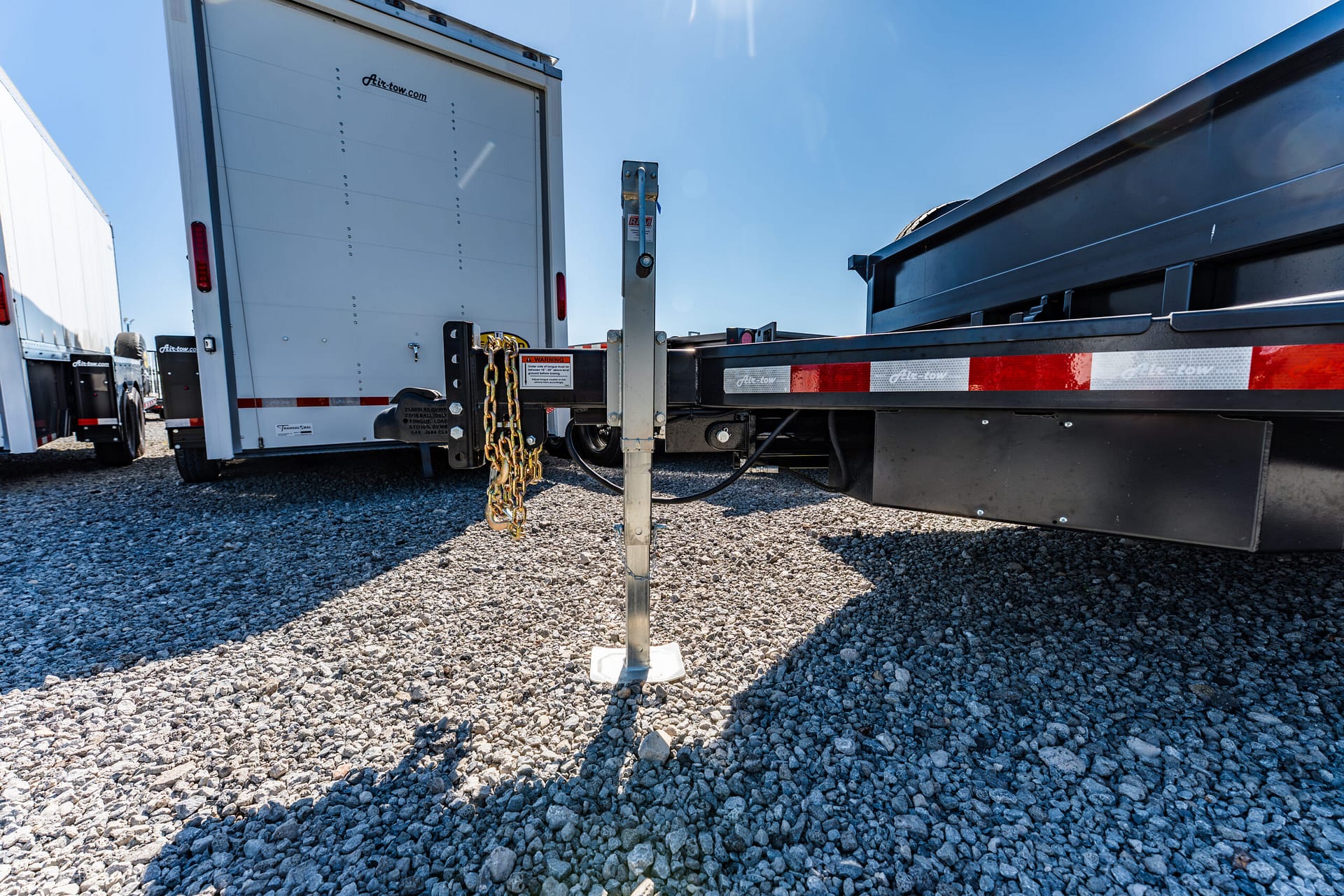 Close-up of a trailer hitch with safety chains hanging, connected to a gooseneck ball on a gravel lot, with a white trailer visible in the background and blue sky above.