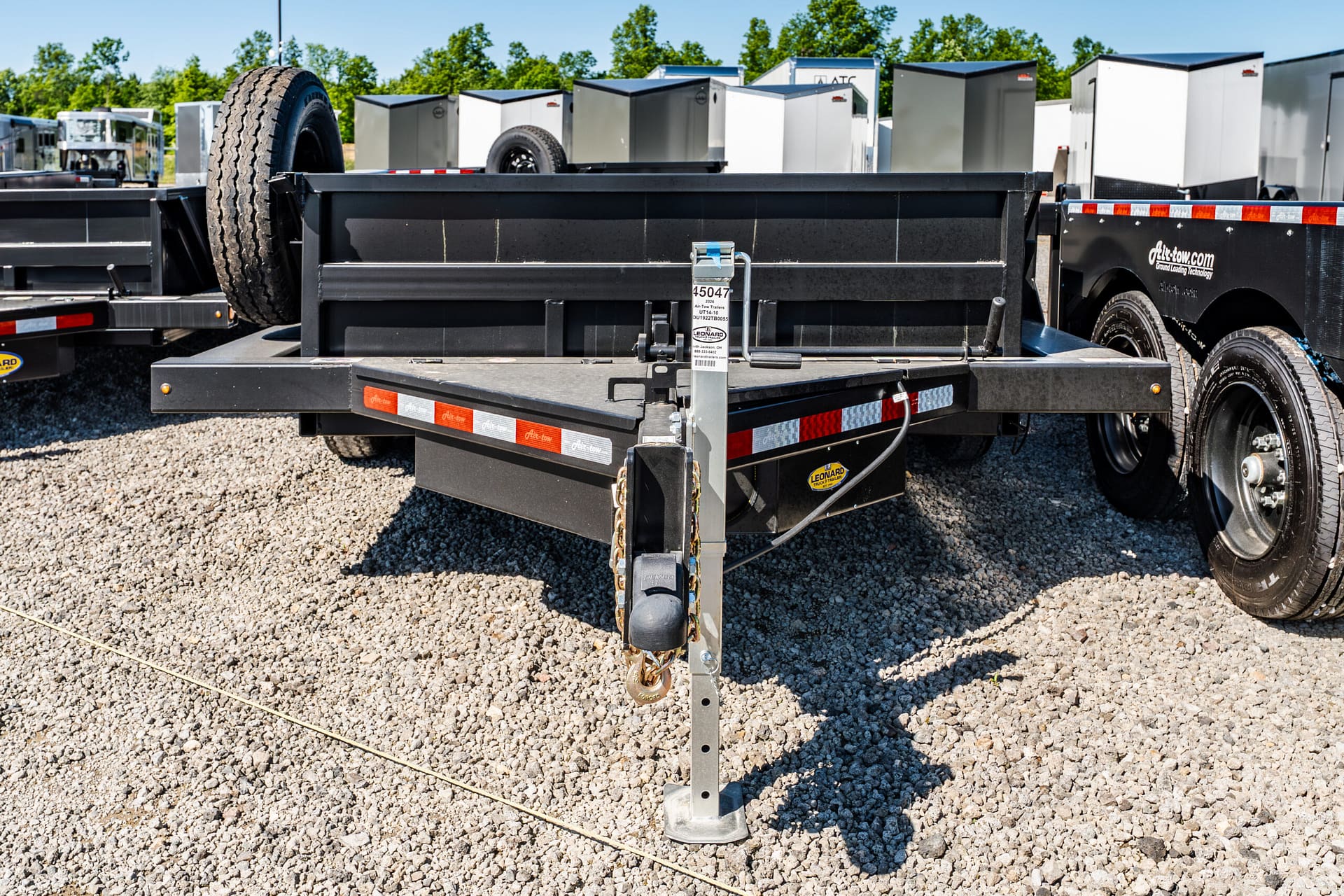 Front view of a black flatbed trailer hitch on a gravel lot, with a safety chain and a mounted spare tire, and other trailers in the background.