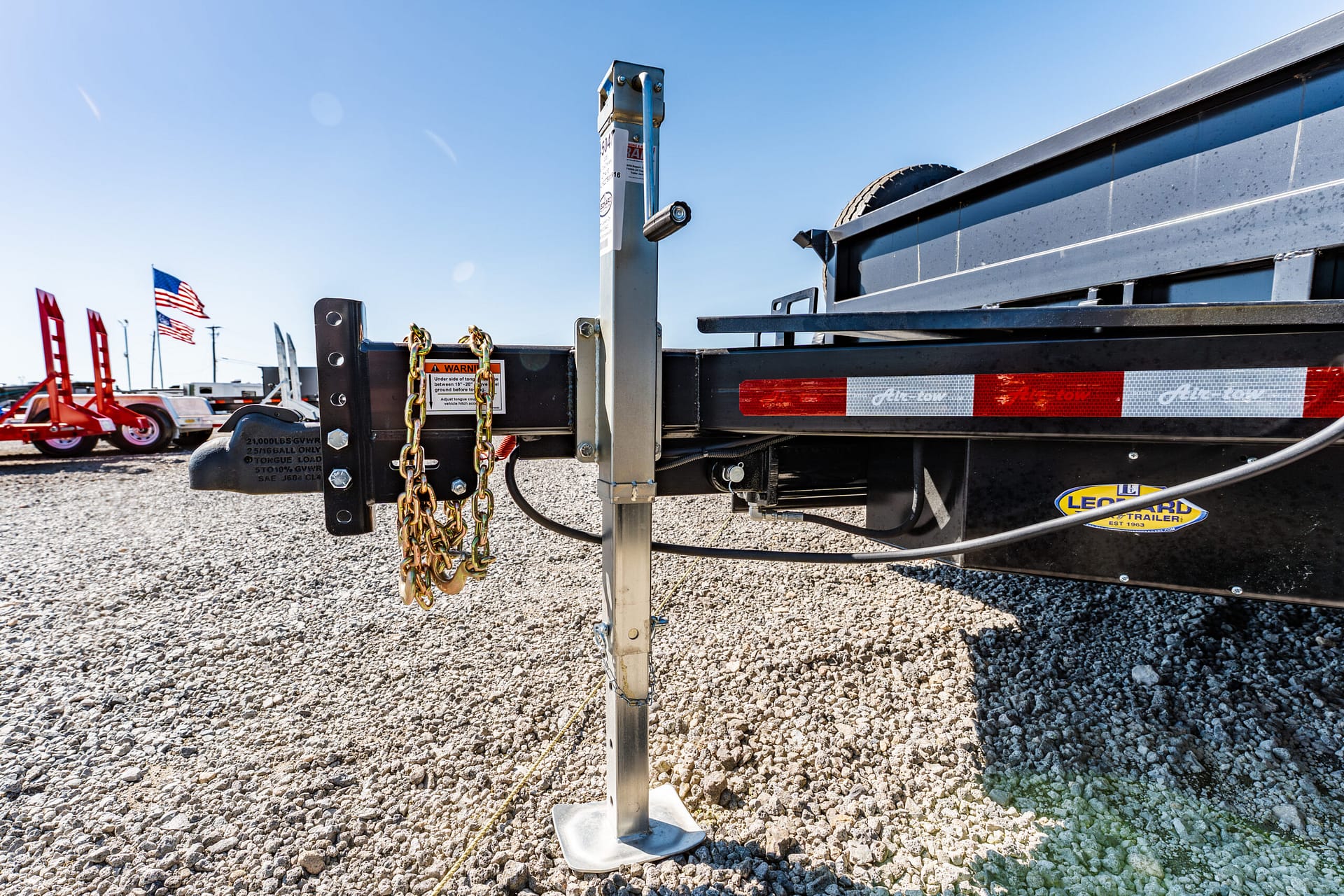 Close-up of a trailer hitch with safety chains hanging, metal frame, and a gravel lot under a clear blue sky; flags visible in the background.