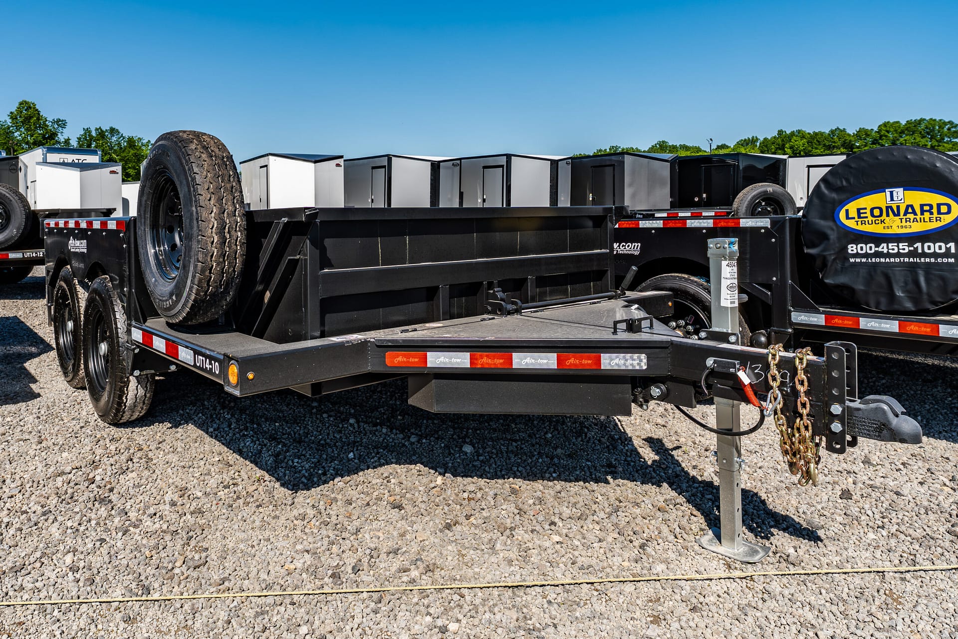 Flatbed trailer with a large spare tire and safety chains, logoed Leonard Trailer in a gravel lot with other trailers behind it.