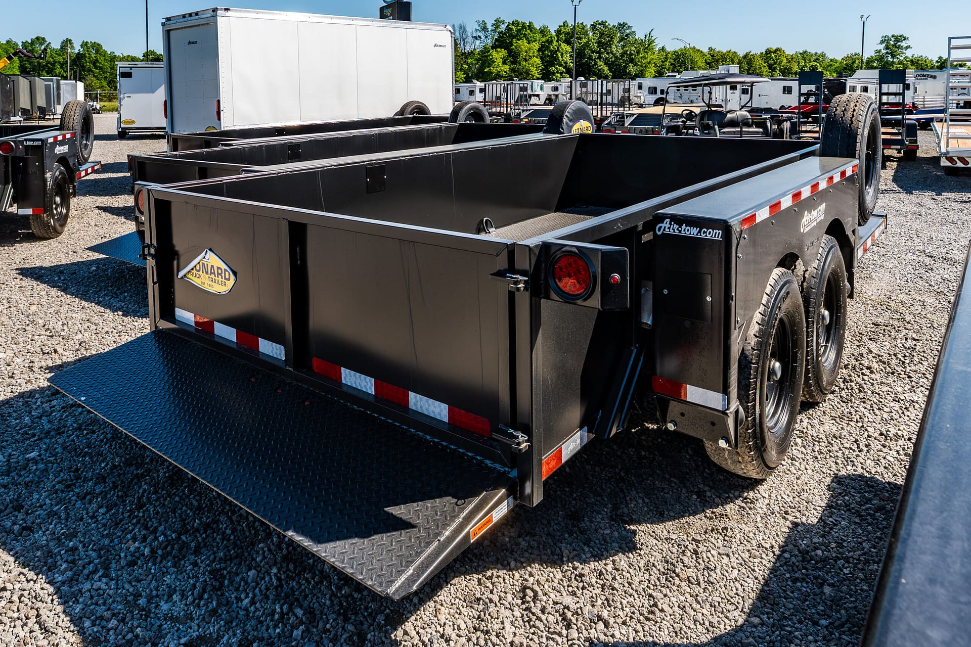 Black flatbed trailer with a diamond-plate ramp in the foreground, parked on a gravel lot with other trailers and trailers’ tires visible in the background.
