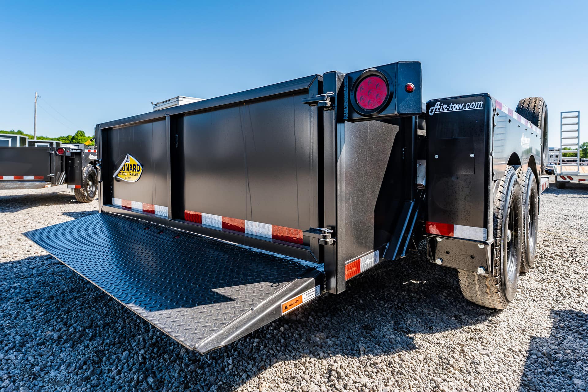 Black dump trailer with a lowered diamond-plate ramp on a gravel lot under a clear blue sky, showing a red taillight at the rear side.