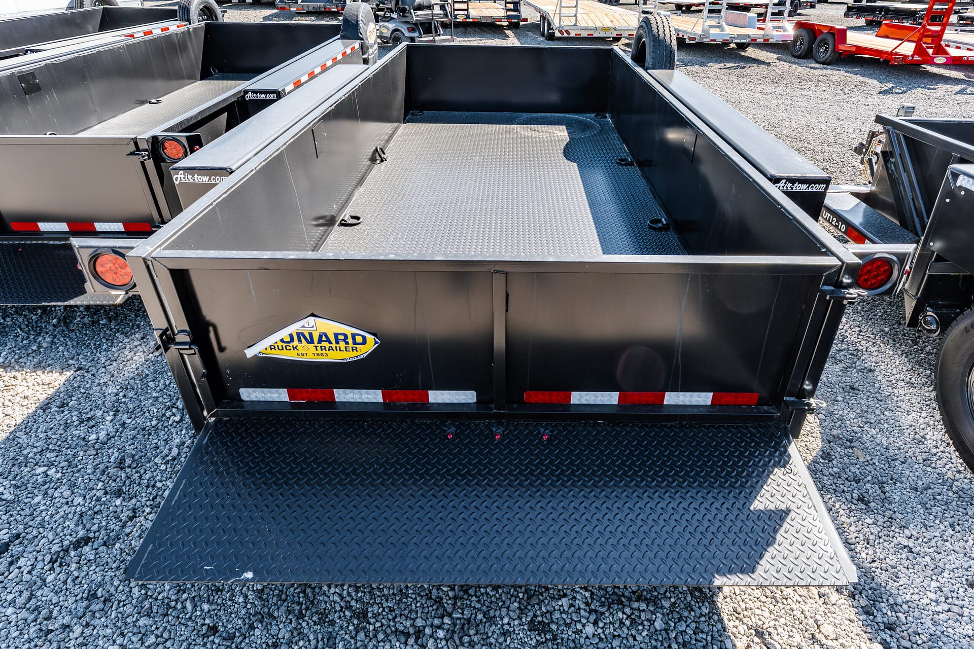 Open flatbed trailer with a diamond-plate steel floor and black side walls, parked in a gravel lot with other trailers nearby.