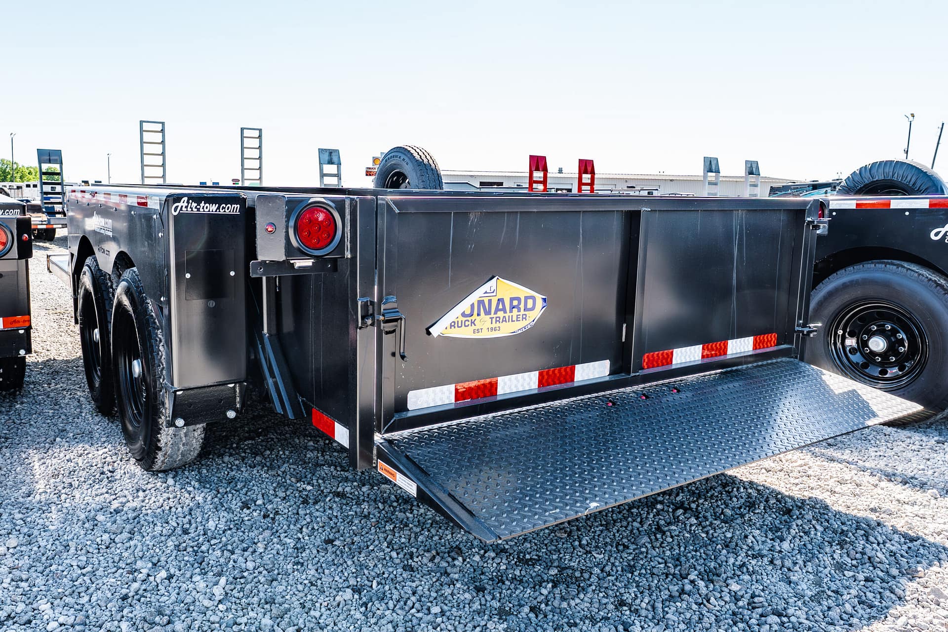 Black flatbed trailer with a lowered diamond-plate ramp on a gravel lot; red taillights visible at the back.