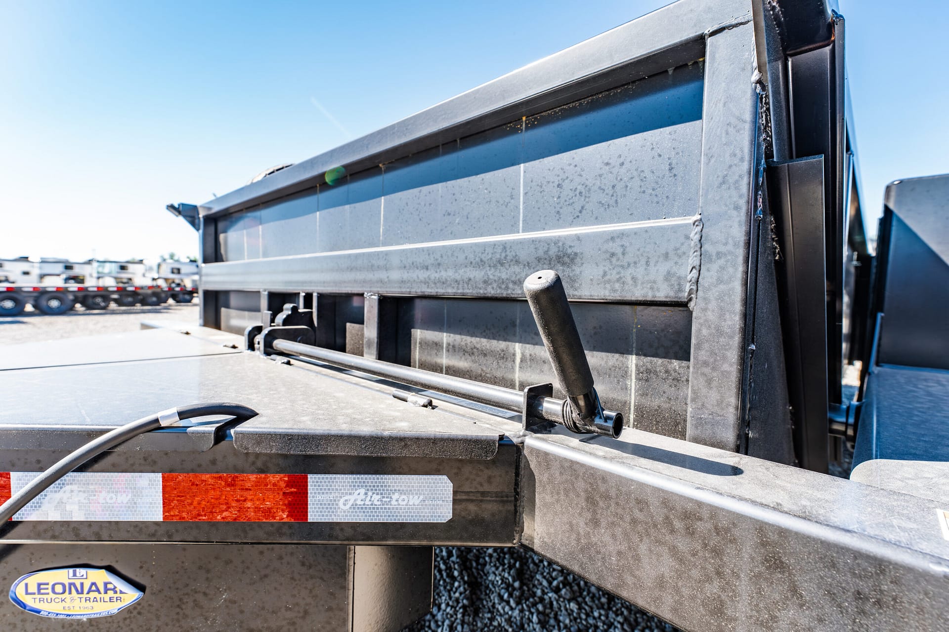 Close-up of a dump truck bed with a metal side panel, hydraulic lever, and red reflector strip under a clear blue sky.
