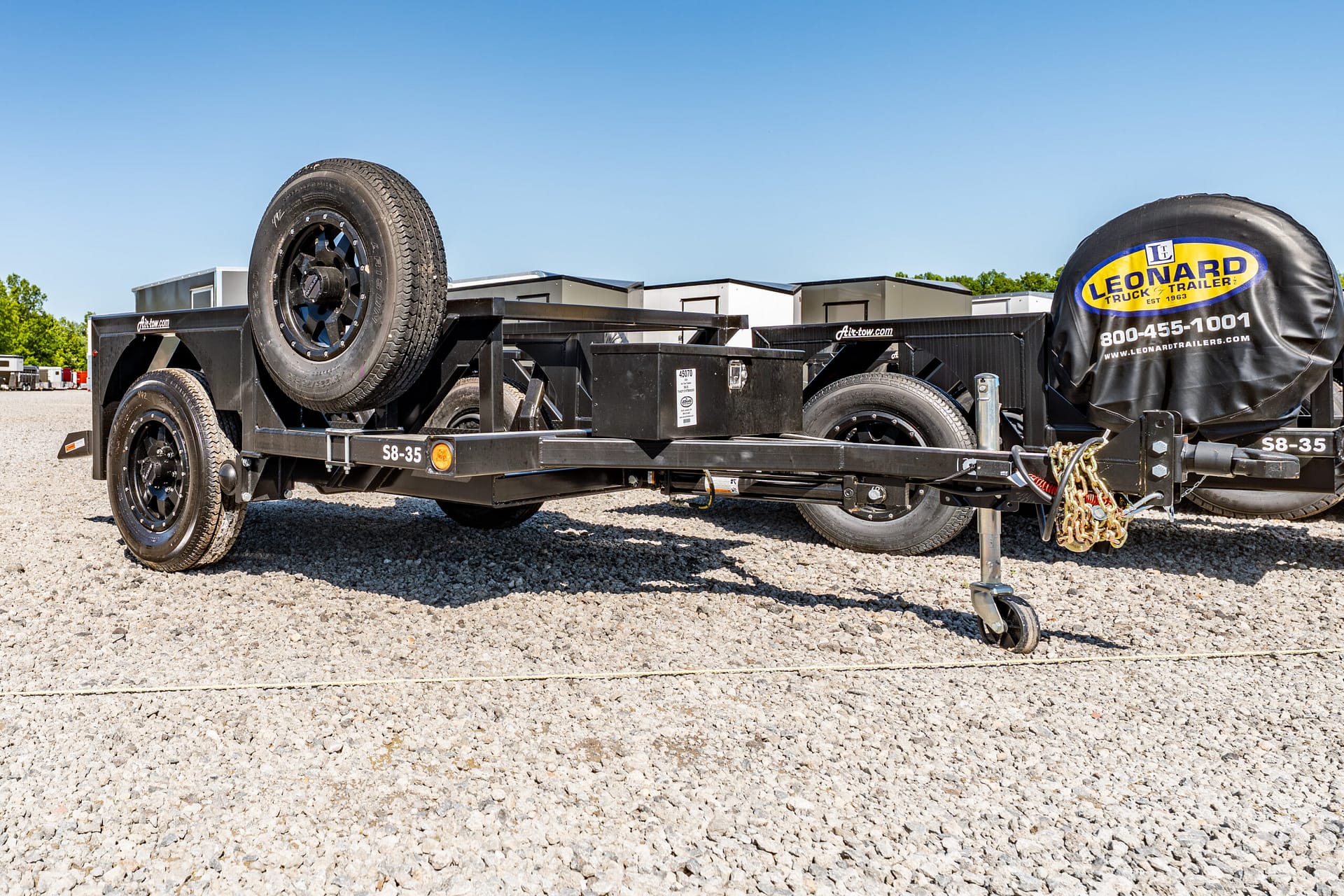 Black utility trailer on a gravel lot with a mounted spare tire on the front corner and a blue sky overhead
