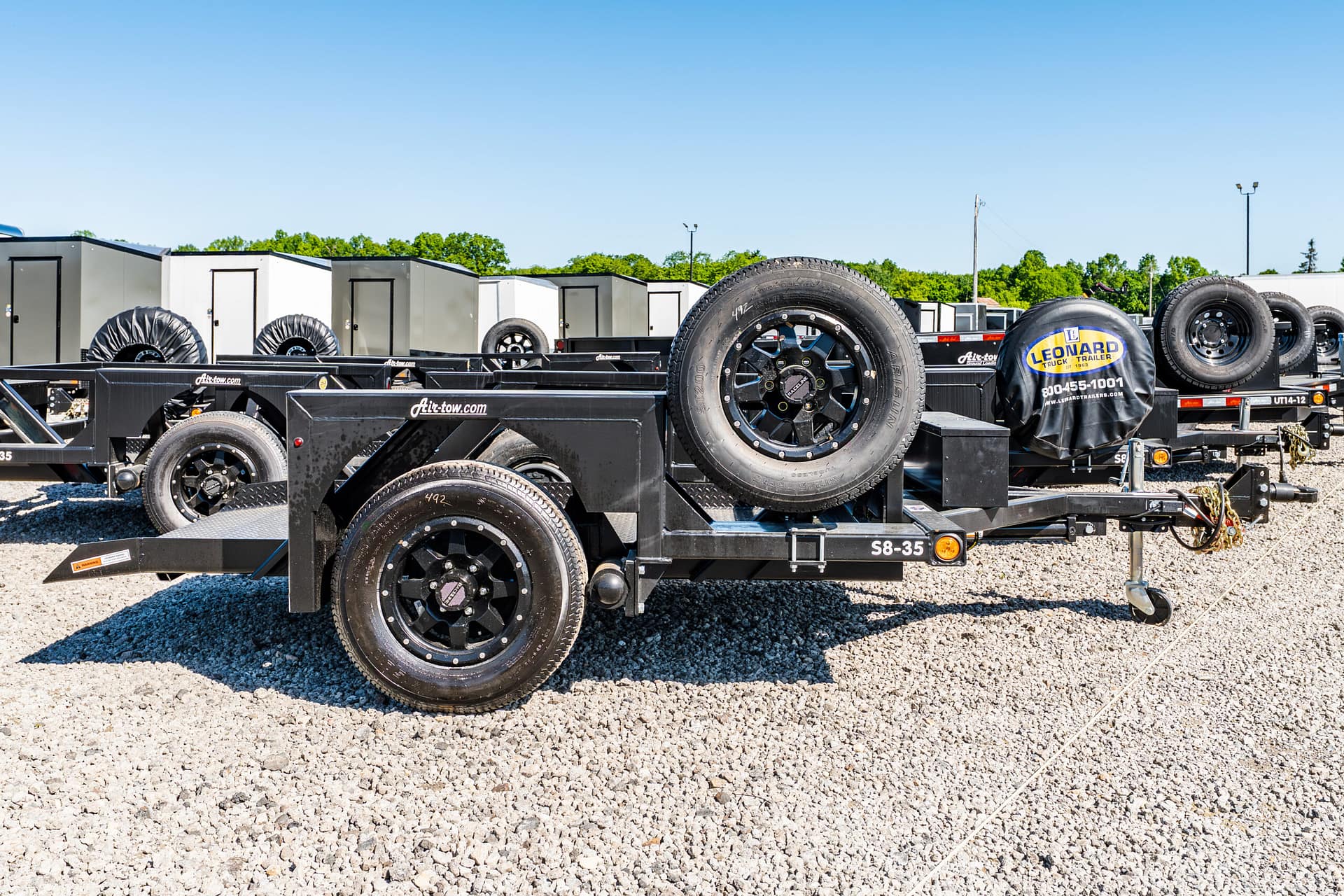 Row of black trailer frames with mounted spare tires in a gravel lot under a clear blue sky.