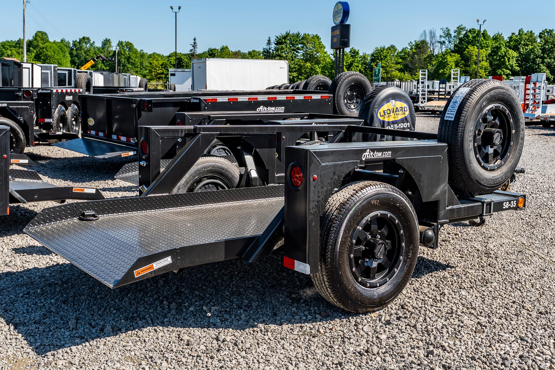 Black multi-axle trailer frames with spare tires in a gravel lot, rows of equipment trailers under a clear blue sky with trees in the background.