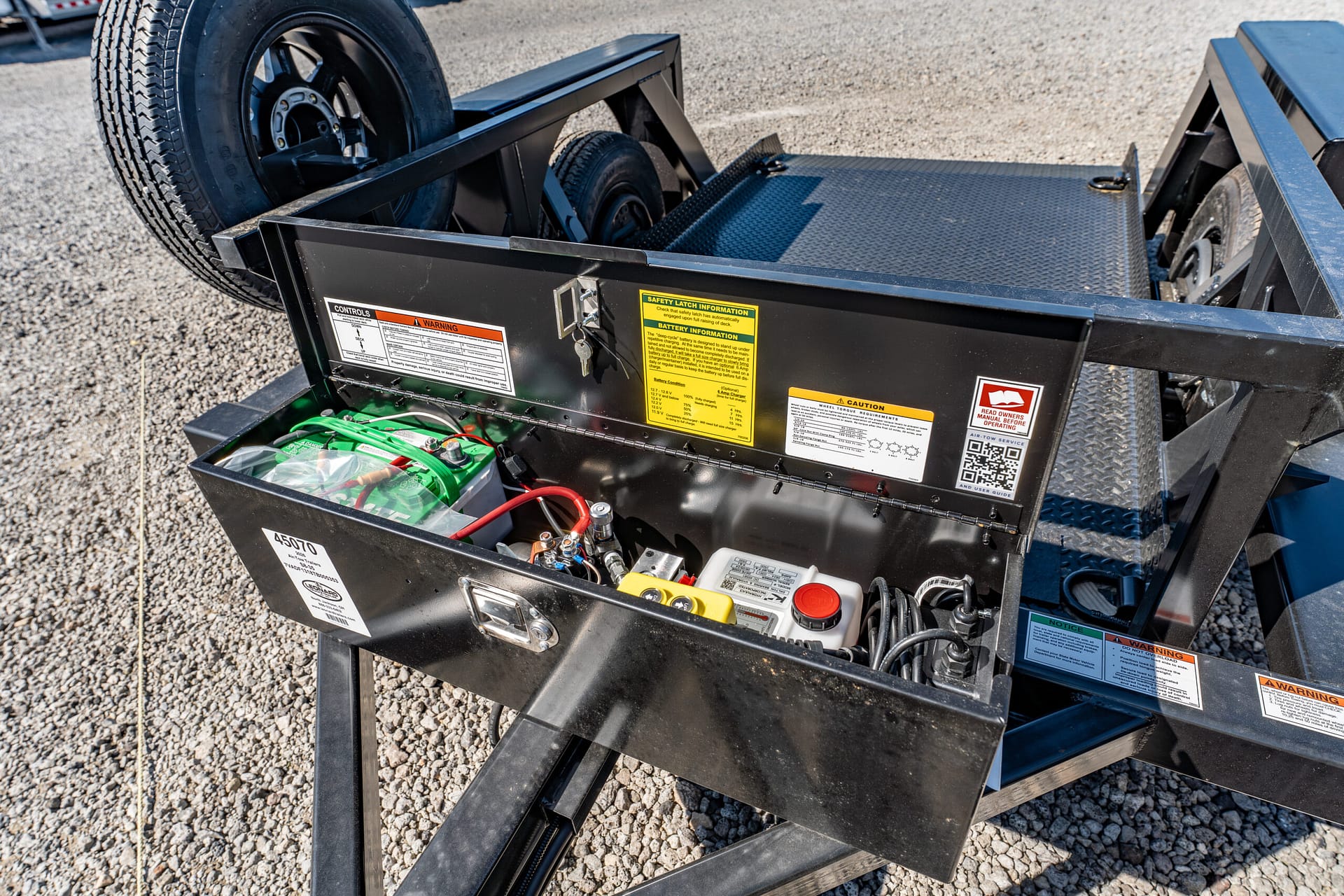 Open compartment of a black trailer showing a battery, wiring, and warning labels inside the toolbox area