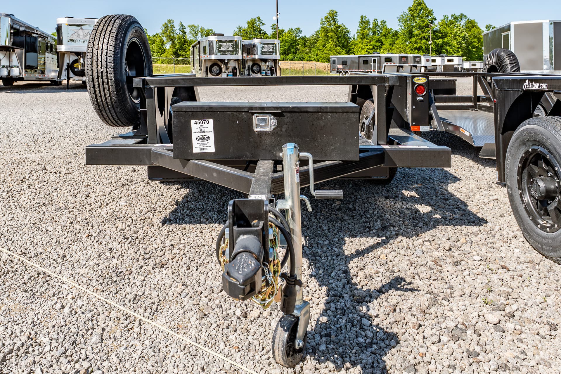 Close-up of a black trailer hitch and spare tire on a gravel lot, with other trailers in the background.