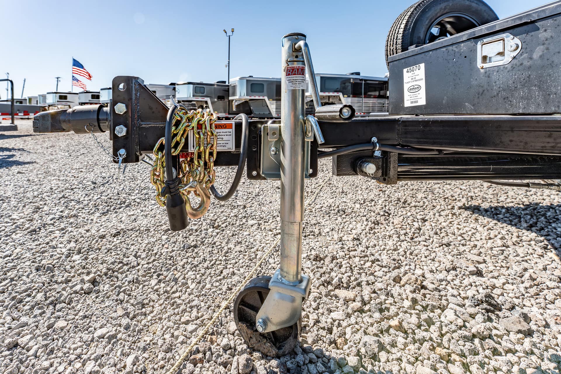Close-up of a trailer hitch with safety chains and a wheel jack on a gravel lot, RVs in the background and an American flag in view.