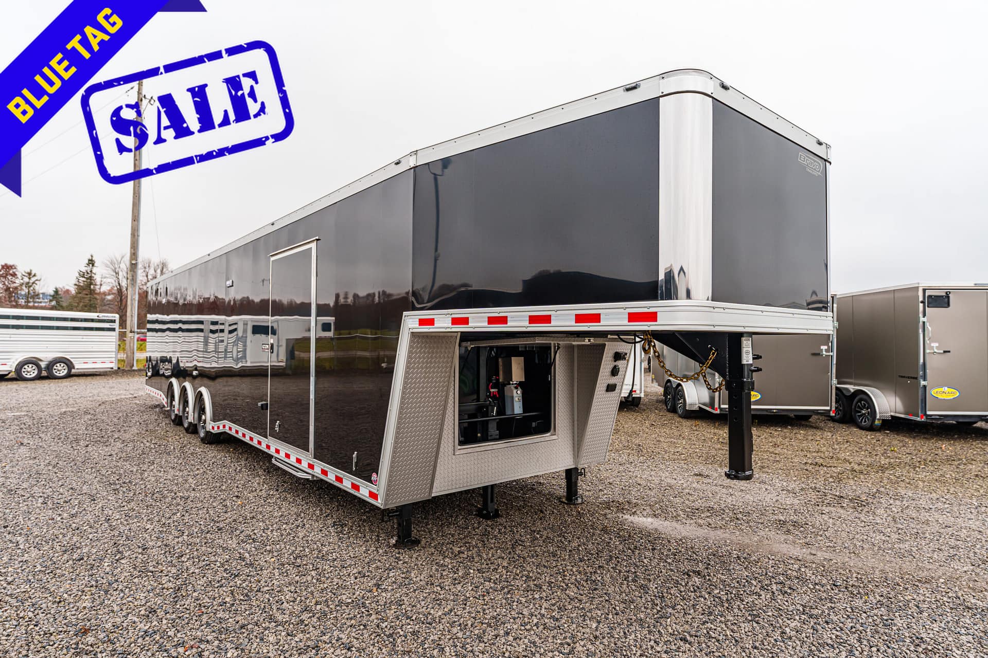 Large black trailer on a gravel lot with a blue 'Blue Tag' sale ribbon in the corner and the rear door open to reveal equipment inside.