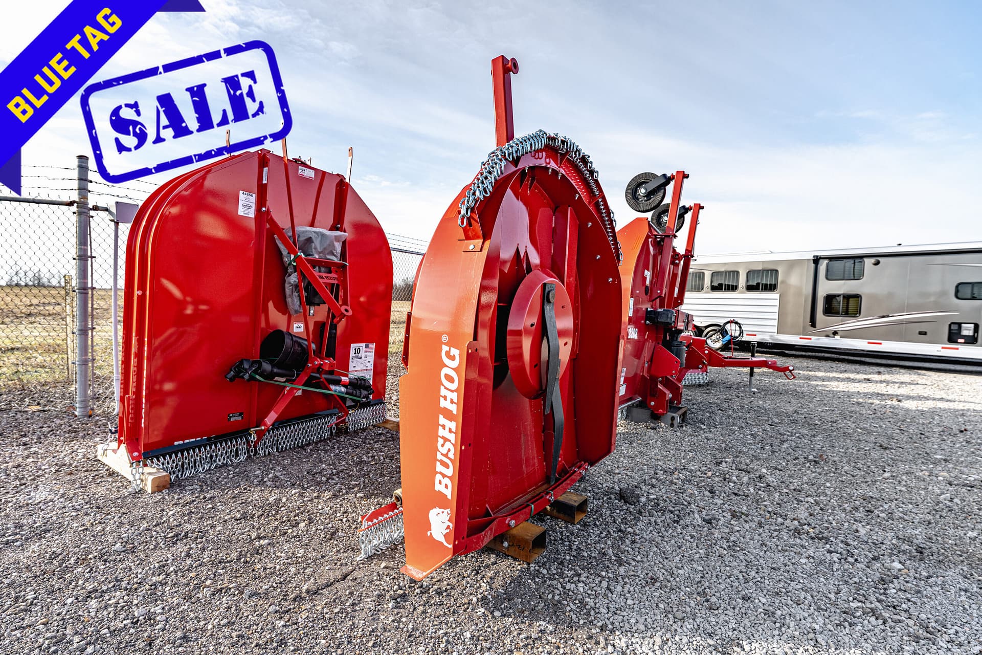 Red industrial farm equipment on a gravel lot with a blue 'Blue Tag Sale' banner in the corner and a trailer in the background
